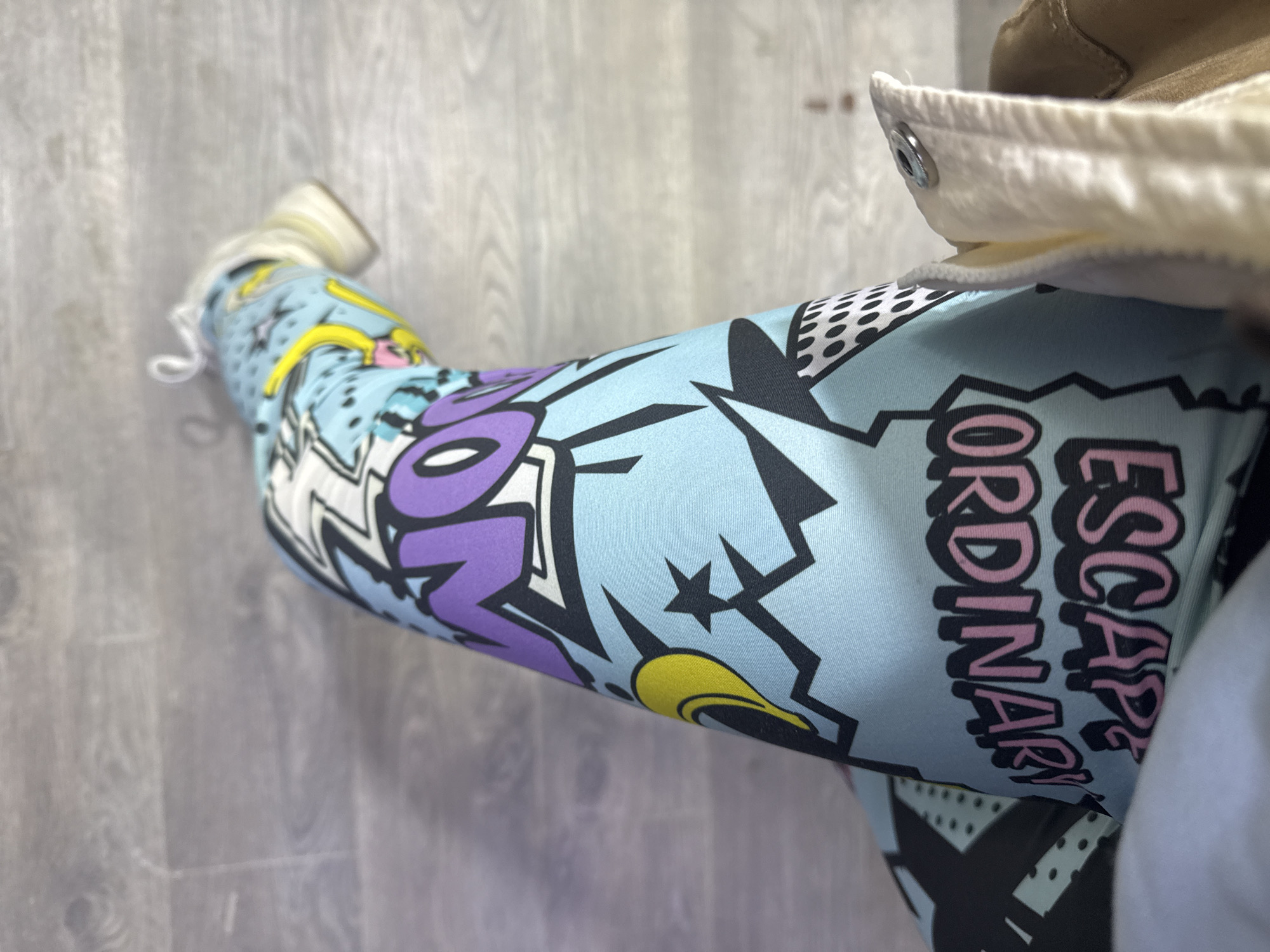 A close-up, angled shot shows a person wearing comic-style patterned leggings with a sneaker visible at the far end of the leg. The leggings feature bold graphics including purple lettering, black shapes, yellow accents, and the phrase "ESCAPE ORDINARY" in pink. A jacket or garment with a white, quilted lining and a metal snap is visible overlapping the top of the leggings near the right edge of the image. The floor is a light grey with a wood-grain pattern. The location is not discernible from the image.