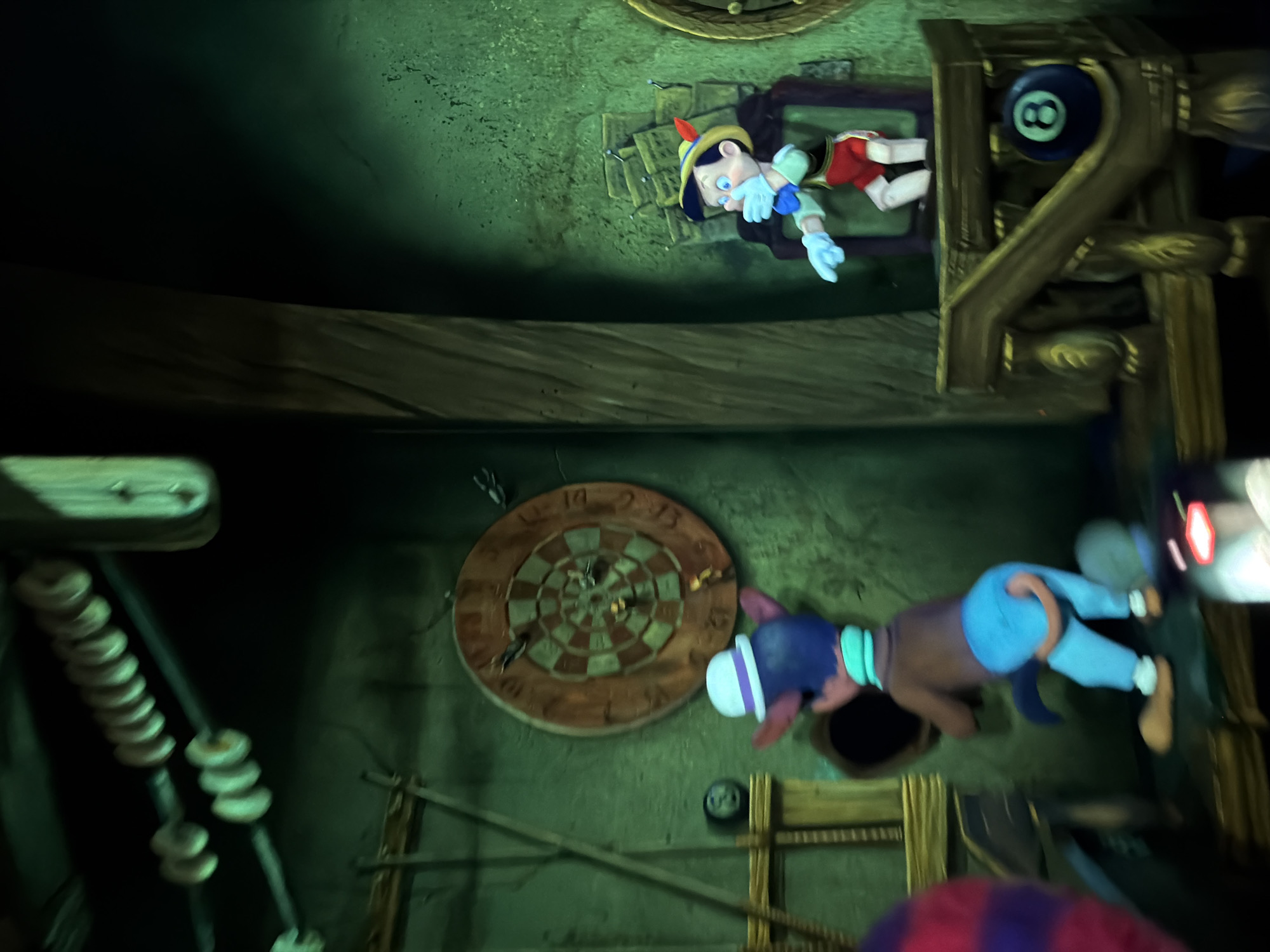 The image captures a scene from the dark ride attraction "Pinocchio's Daring Journey" located in Disneyland Park in Anaheim, California, United States of America. In the foreground, a costumed character resembling Gideon the Cat from the Disney film *Pinocchio* is seen from behind. He is wearing a white hat with a purple band, a teal scarf, and bright blue pants, with his back turned to the viewer. He appears to be interacting with a dark opening in the wall. To the right, Pinocchio, the wooden puppet, is depicted in his iconic outfit of red shorts, a yellow hat with a red feather, and blue gloves. He is shown with his hand over his mouth, a common animated expression. Behind Pinocchio, several framed pictures, possibly a part of the attraction's set design, are visible. A dartboard hangs on the wall between the two characters, with several darts stuck in it. To the left of the dartboard, an eight-ball from a pool game is placed on a wooden shelf. The environment is dimly lit and has a rustic, somewhat spooky atmosphere, characterized by dark green walls and wooden structures.
