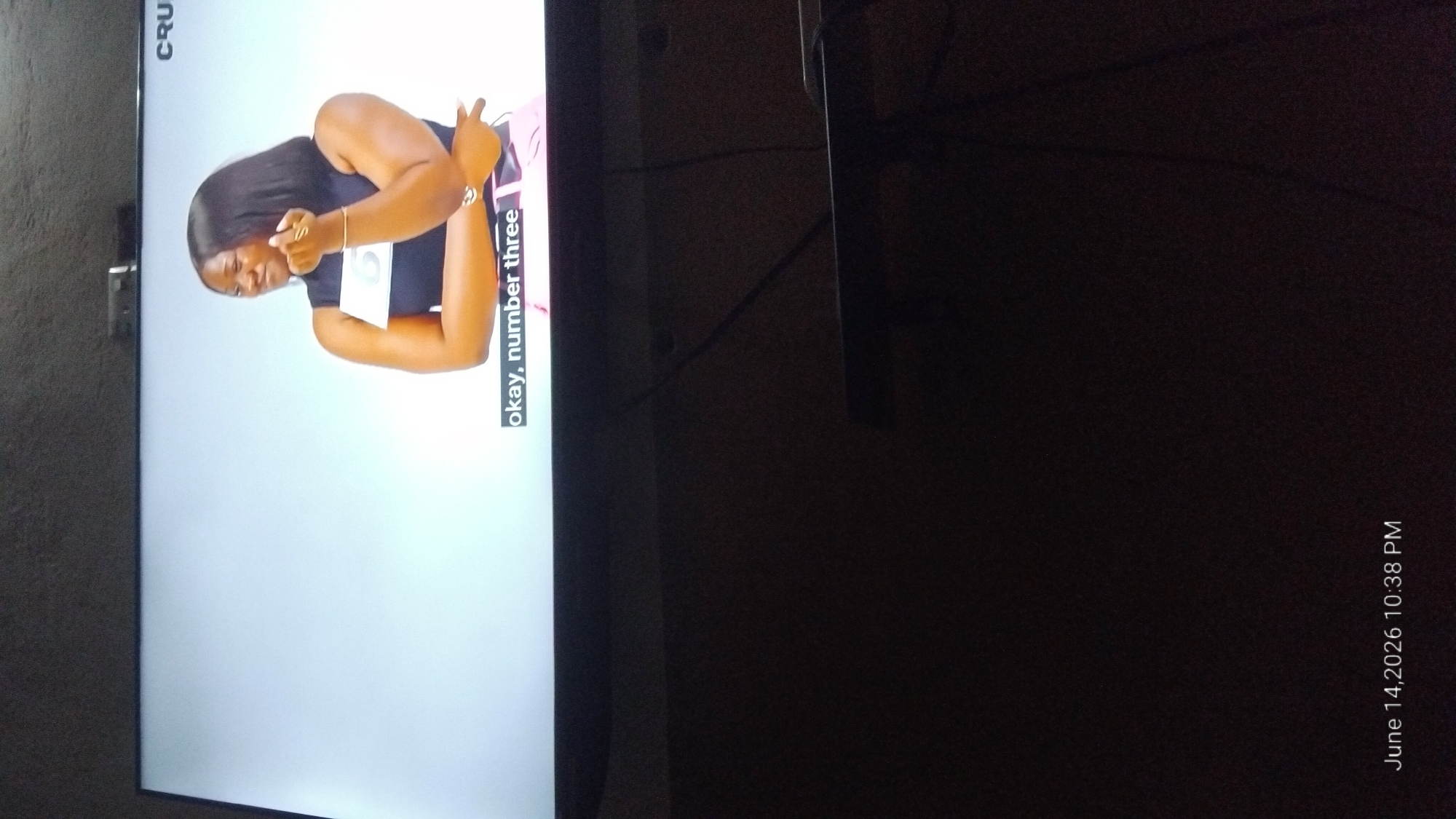 A young Black woman with dark hair is on a television screen, wearing a black sleeveless top and pink bottoms. A white tag with the number "6" is attached to her top. She is looking to her left, with her right hand raised and touching her chin. Subtitles at the bottom of the screen read "okay, number three." The date and time stamp "June 14, 2026 10:38 PM" is visible in the lower left corner. The television is dark, suggesting the room is dimly lit.