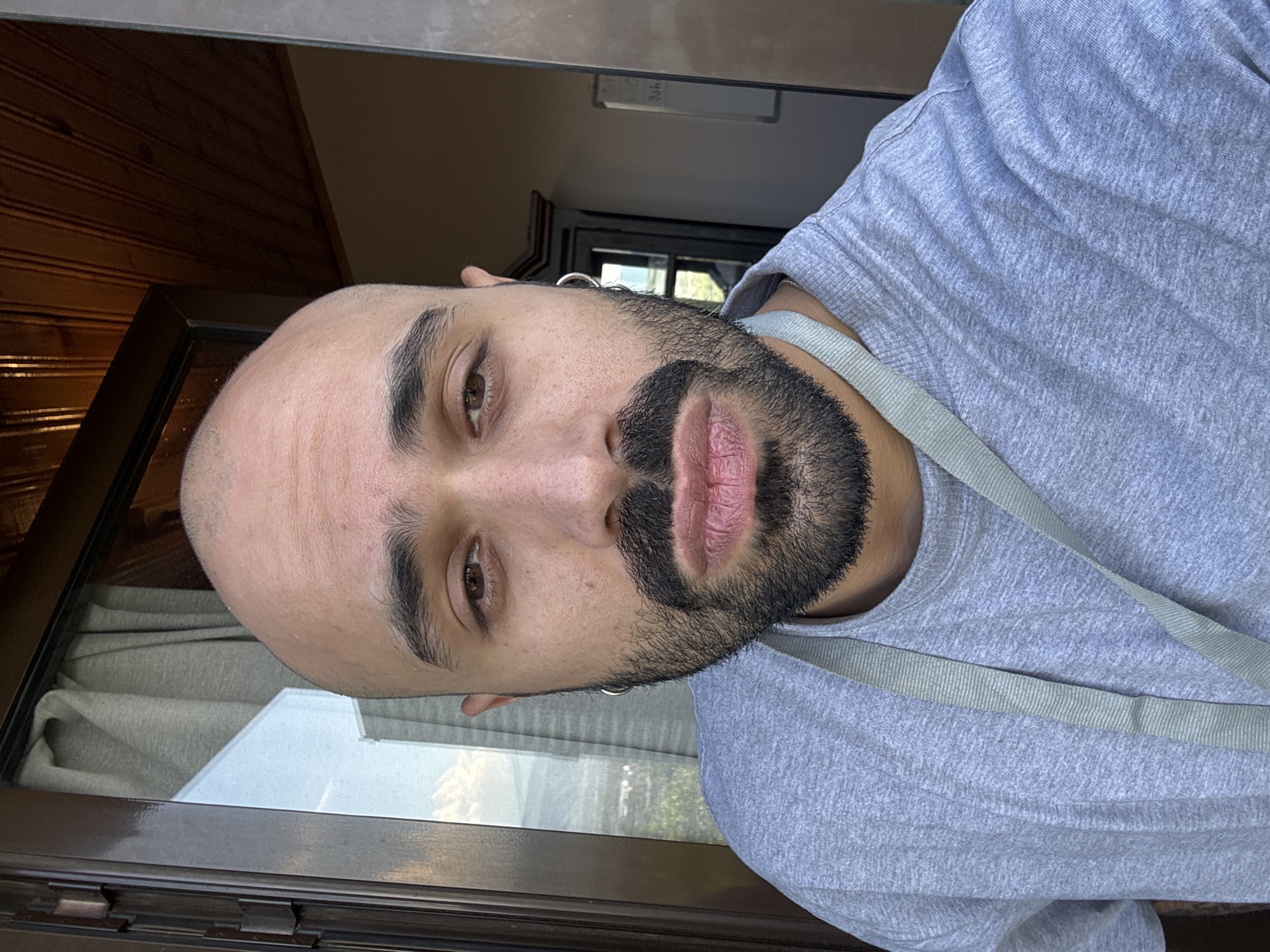 The image is a rotated close-up selfie of a man with a shaved head, dark beard, and mustache. He has brown eyes and thick eyebrows, looking directly at the camera with a neutral expression. A silver hoop earring is visible in his left ear. He wears a light grey short-sleeved t-shirt and a light grey strap across his right shoulder. His lips appear somewhat dry or chapped.
The background shows an indoor setting with dark wooden paneling on one side. Behind him is a dark-framed window or glass door, through which a blurred outdoor scene with greenery and distant elements is visible, indicating it is daytime. On the window frame, a small, white rectangular label with faint, almost illegible text (possibly "0.4h") is present. The scene is located in Mtskheta, Georgia.