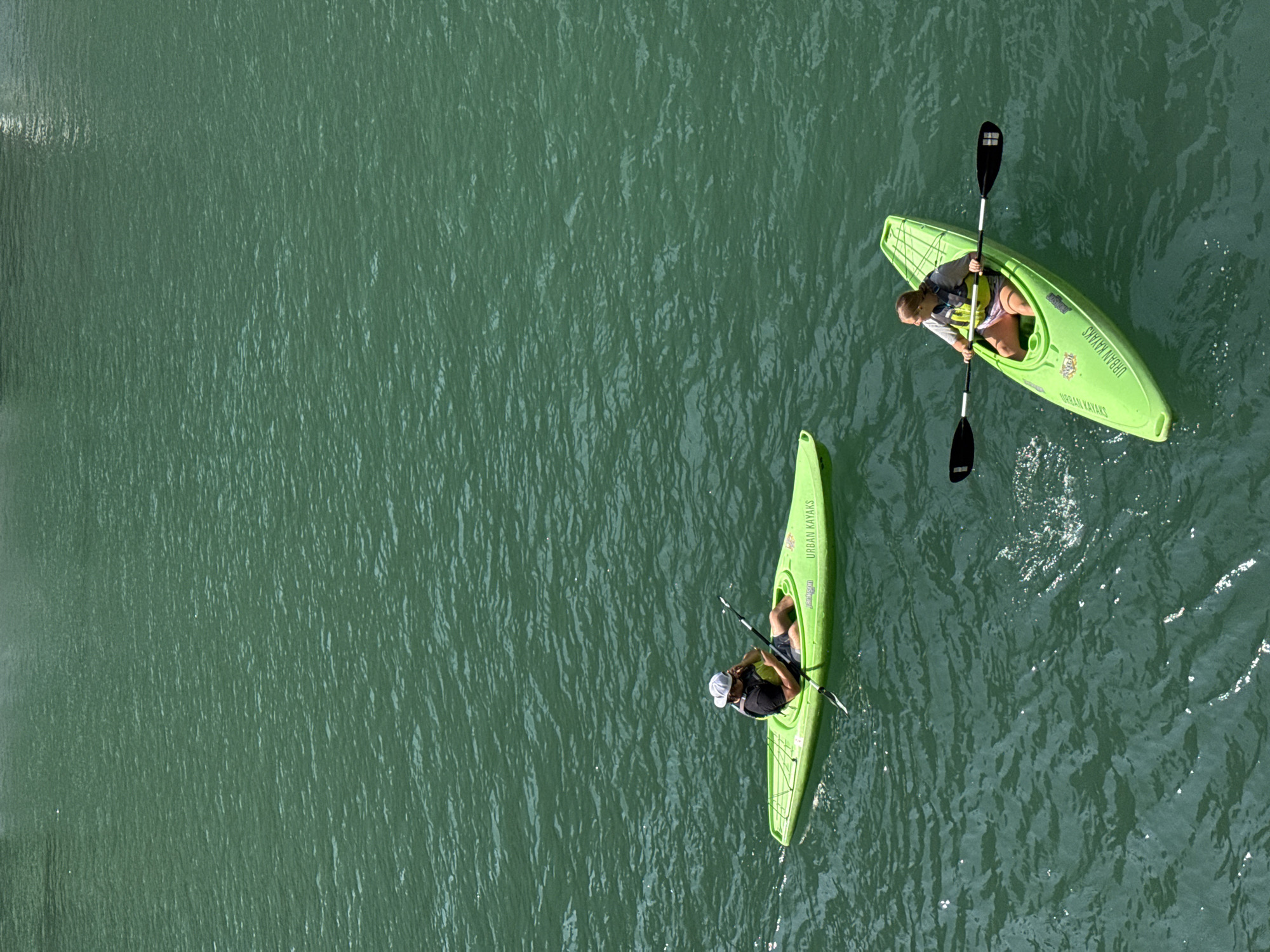 Two people are kayaking on a green body of water. The person in the foreground is a woman wearing a life vest and shorts, paddling a bright green kayak. The kayak has "URBAN KAYAKS" printed on its side. The person in the background is a man wearing a life vest and a hat, also in a bright green kayak, holding a phone to his ear. The water appears calm with subtle ripples. The image is taken from an elevated perspective. The location is likely a navigable waterway within the city of Chicago, possibly a river or canal, given the presence of "URBAN KAYAKS."