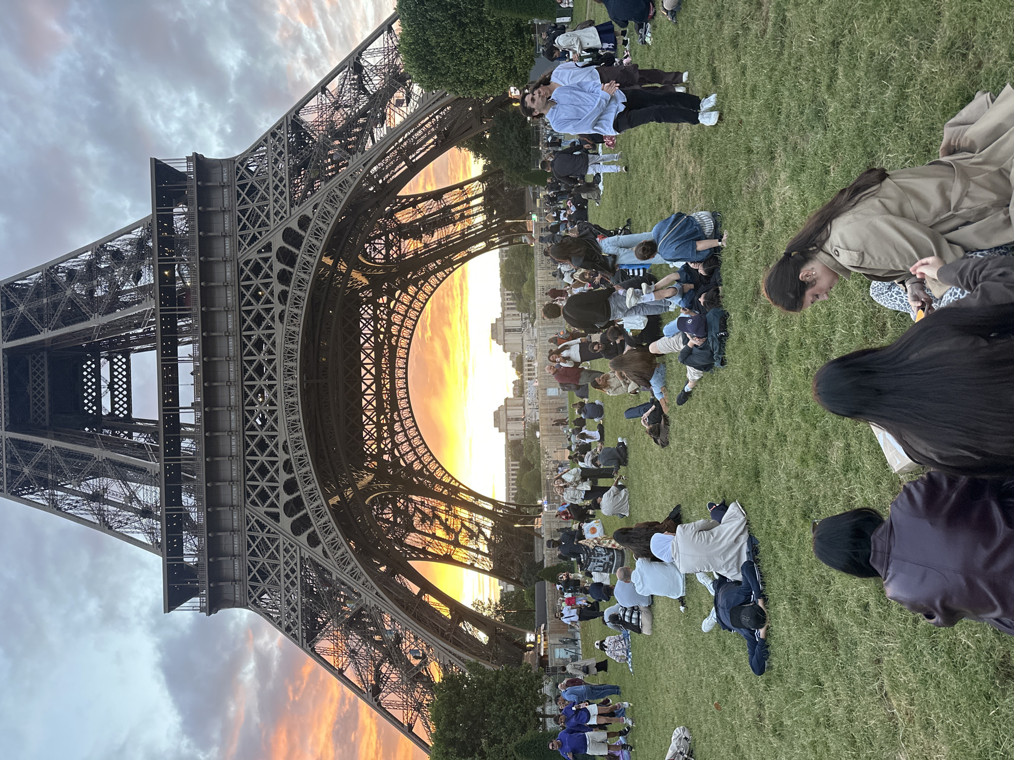 A picturesque scene captures the iconic Eiffel Tower in Paris, France, during sunset. The image, presented with a sideways orientation, features the colossal iron lattice structure of the Eiffel Tower dominating the left side of the frame. The setting sun casts a radiant orange and yellow glow, visible directly through the tower's lower arch, creating a stunning silhouette effect and illuminating the intricate metalwork. The sky above displays a mix of clouds, painted with soft hues of grey, pink, and orange, characteristic of twilight.
On the expansive grassy area of the Champ de Mars, numerous people are gathered. Most are seated or lying on the green lawn, appearing relaxed as they enjoy the view of the tower and the beautiful sunset. Dressed casually, they form small groups or sit individually, observing the surroundings. The overall atmosphere conveys leisure and enjoyment, typical of visitors at this popular landmark.
Notable details include the names of French engineers and scientists inscribed on the Eiffel Tower's girders, which are visible in parts. These inscriptions include "CAUCHY," "BELGRAND," "FRESNEL," "DE PRONY," "ARAGO," "POISSON," and "MONGE," among others. In the distance, beneath the tower's arch, glimpses of urban buildings can be seen, completing the Parisian vista under the warm, golden hour light.