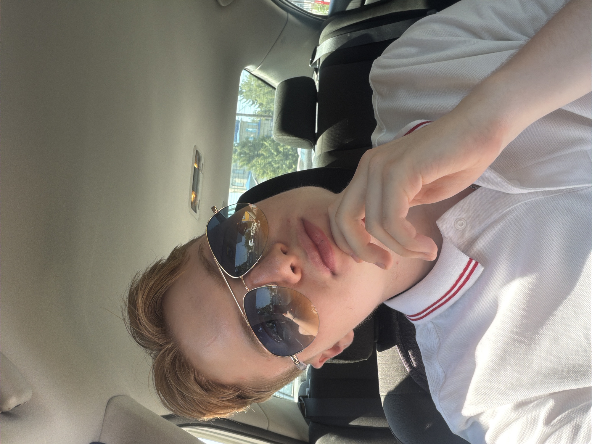 A young Caucasian male with short, reddish-blonde hair and light skin is seated in the back of a car, wearing aviator sunglasses and a white polo shirt with red trim on the collar. His right hand is resting on his chin, and he is looking slightly off-camera. The interior of the car, including the headrests and ceiling with a light fixture, is visible. Outside the car windows, trees and buildings suggest an urban environment, possibly in Polonne, Ukraine. Sunlight is filtering into the car, casting shadows.