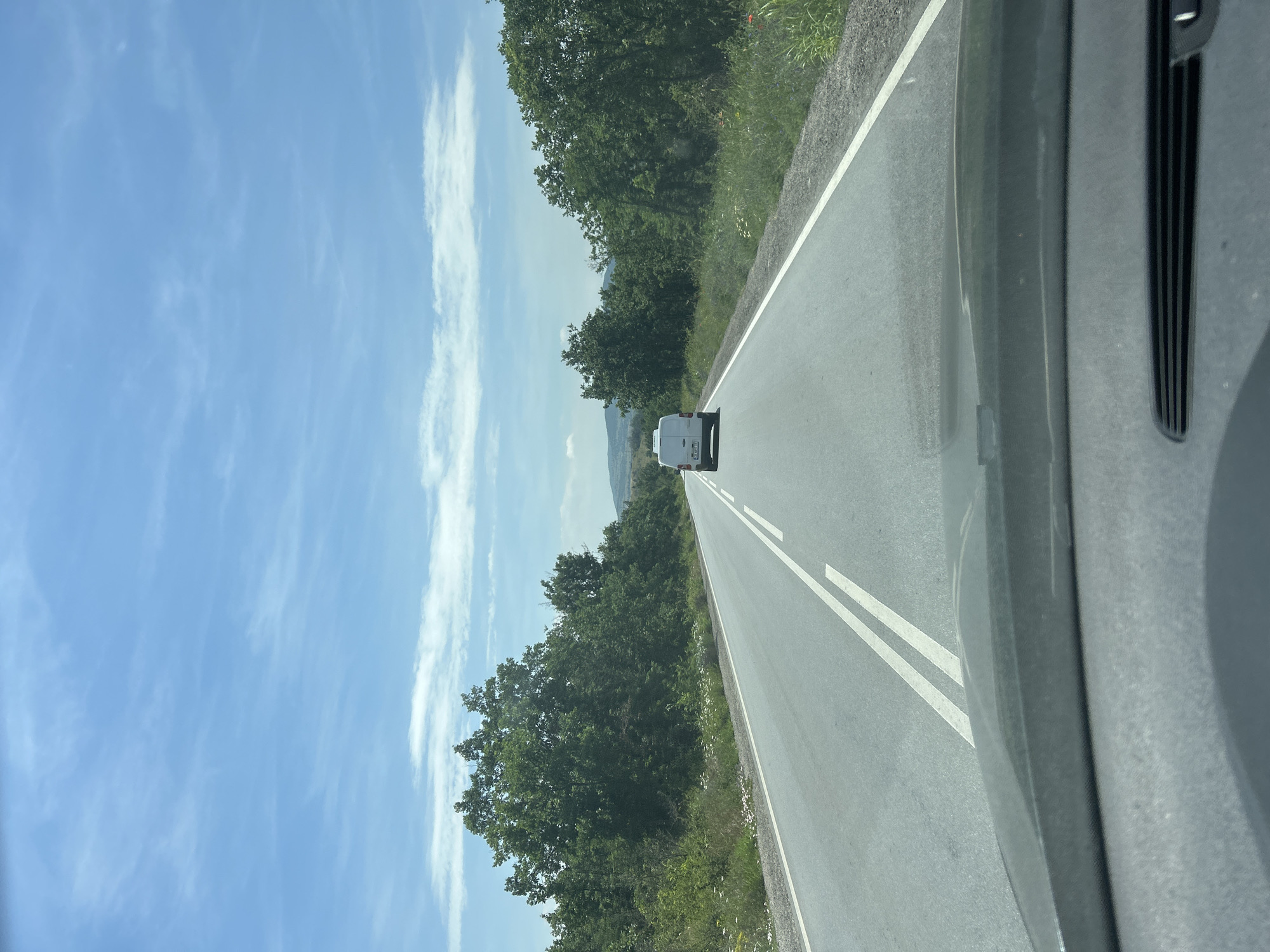 The media file captures a first-person perspective from inside a vehicle, looking out onto an open road in Marino Pole, Bulgaria. The time of day appears to be mid-day, under a bright, clear blue sky adorned with scattered wispy white clouds.
The central element of the scene is a straight asphalt road, receding into the distance. It features double solid white lines in the center, clearly separating lanes of traffic, and a single white line marking the right shoulder. Ahead on the road, a white van is visible, driving in the same direction as the camera. The van's rear license plate, "M 3949 M," is discernible, indicating a Bulgarian registration from the Montana region.
Both sides of the road are bordered by dense green trees and lush vegetation, suggesting a rural or natural setting. Green grass and various wildflowers, including hints of red poppies and white daisies, are visible along the roadside. In the far distance, beyond the trees, the faint outlines of hills or mountains can be seen merging with the horizon.
A portion of the camera's originating vehicle, specifically a dark grey dashboard or pillar, is visible on the right side of the frame, providing context for the view from inside a car. The overall impression is one of a scenic and peaceful drive through a verdant landscape in rural Bulgaria on a clear, sunny day.