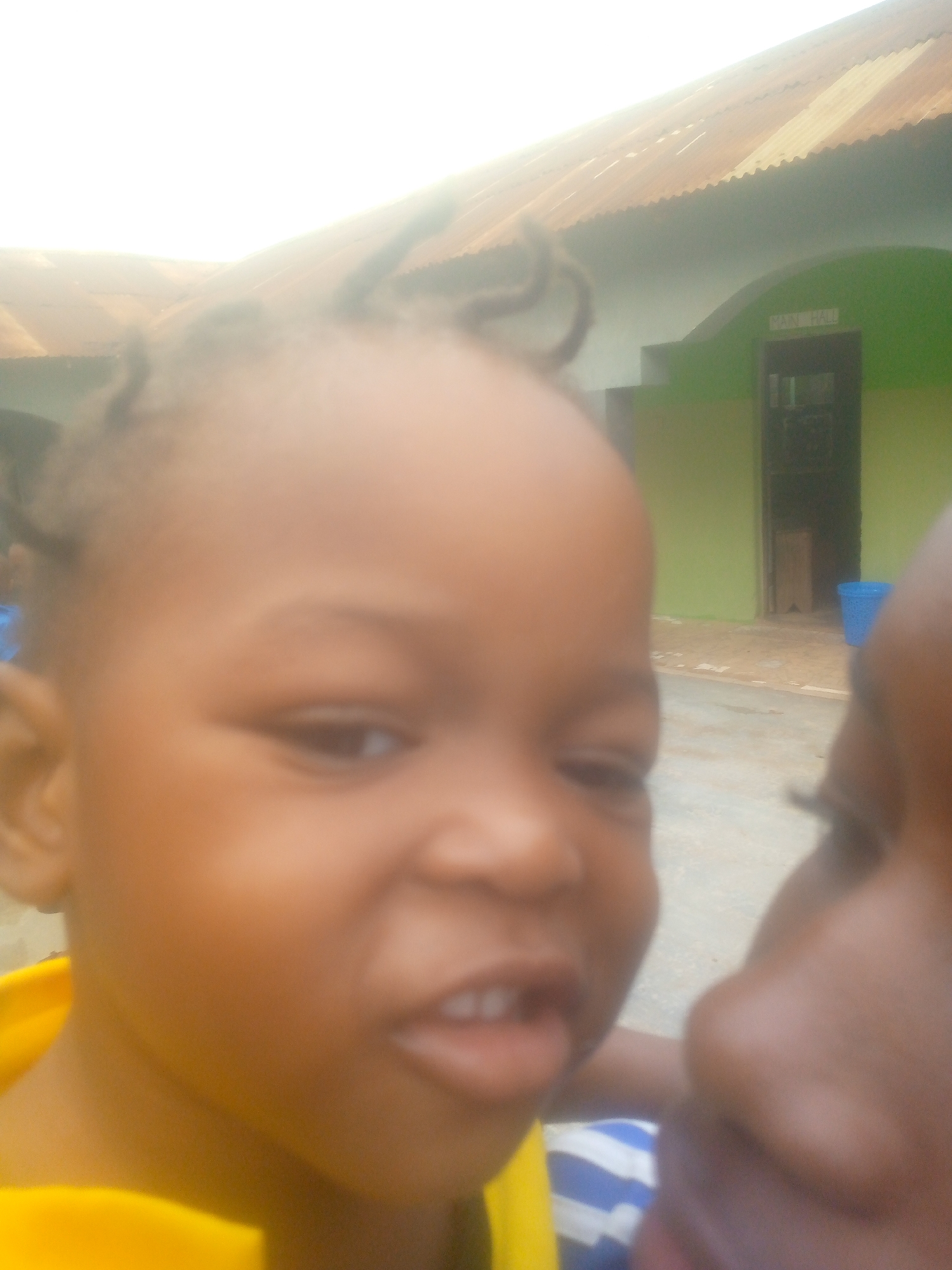 A close-up shot focuses on a child with dark brown skin, short, curly hair, and protruding front teeth. The child is wearing a yellow shirt. To the right of the child, partially out of focus, is another person's face, revealing a dark brown skin tone and a blue and white striped shirt.
In the background, there is a building with a corrugated metal roof. The building is painted green, and a doorway is visible, bearing the words "MAIN HALL" above it. A blue bucket sits nearby on the ground, which appears to be a concrete surface.
Based on the prompt, the location is Ikorodu, Nigeria.