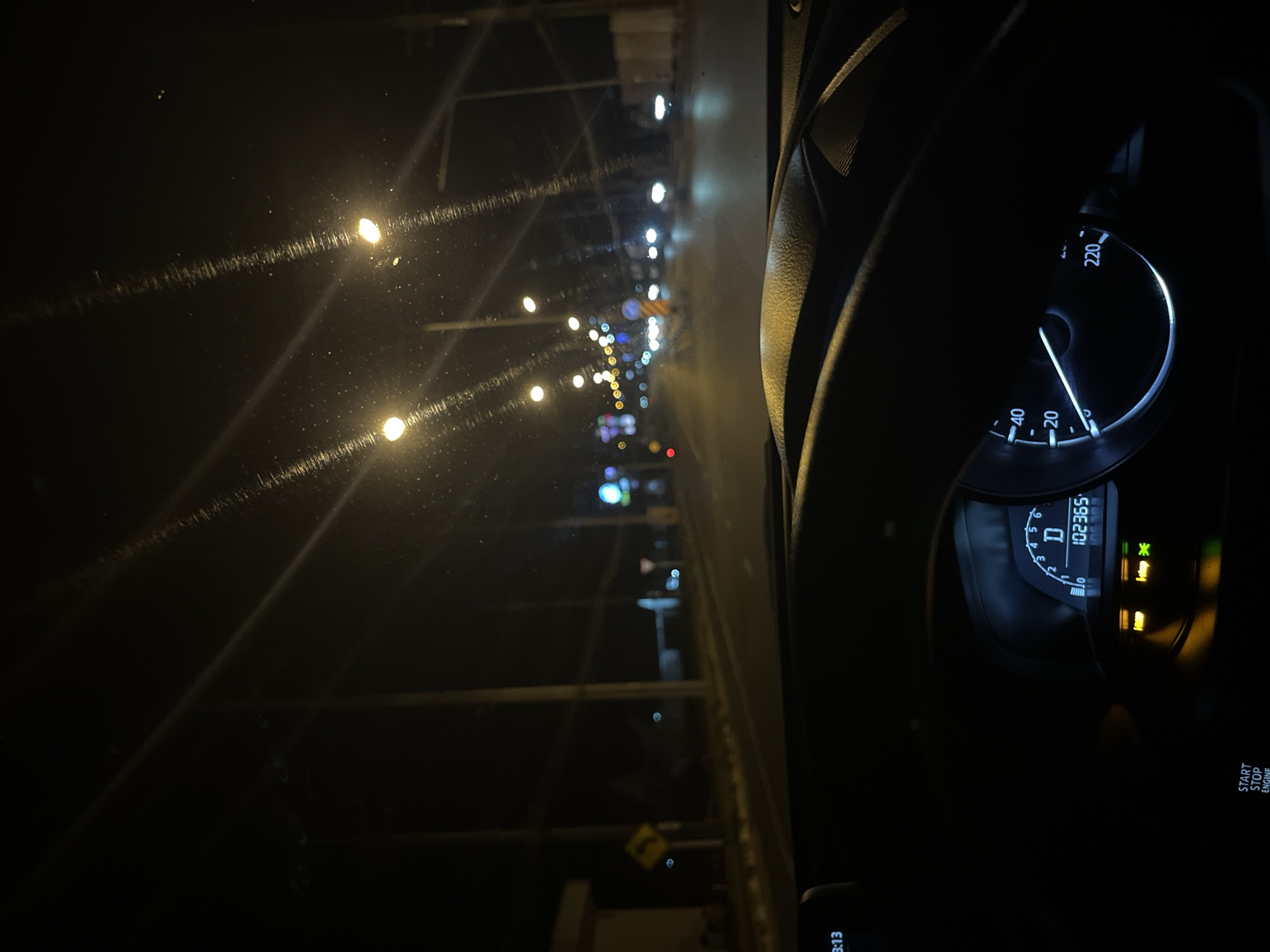 The image captures a nighttime view from inside a car driving on a road in Chalong, Thailand. The foreground is dominated by the dashboard, including a steering wheel and a digital display showing the odometer reading 102365. The dashboard also shows a speed indicator with the needle pointing below 20 km/h. Outside the car, streetlights illuminate the road and the surrounding environment. Traffic lights and illuminated signs are visible in the distance, suggesting an urban setting. The road appears to be paved, and there are utility poles lining the street. The scene is characterized by the darkness of night, punctuated by artificial lighting.