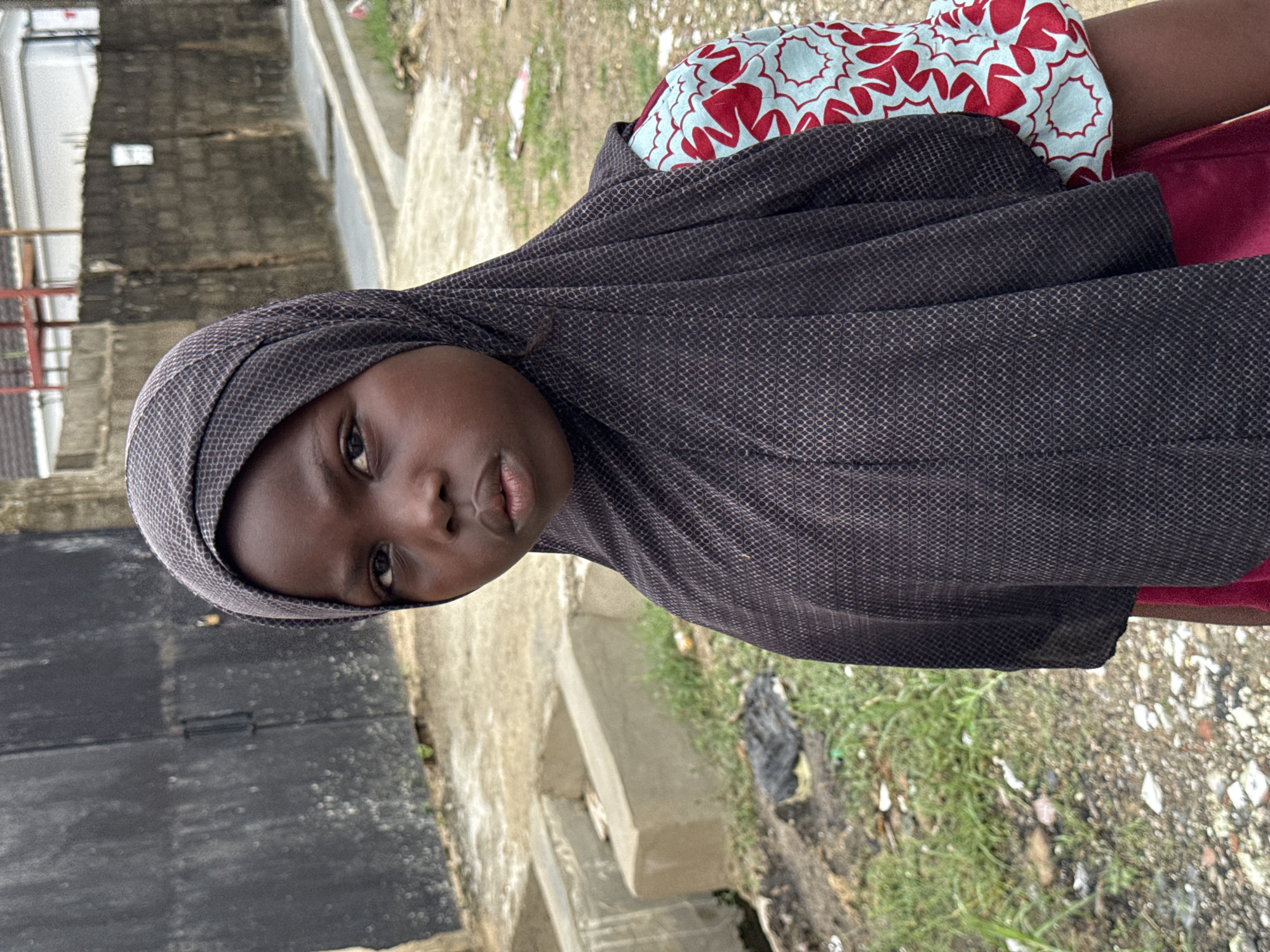 A close-up, medium shot shows a young girl, likely under 12 years old, wearing a dark gray headscarf and facing the camera. Her expression is neutral. Visible behind the headscarf are portions of her clothing: red and a patterned light blue and red garment. The background is an urban environment, appearing to be in a state of disrepair. To the left is a dark gray or black wall or fence. Below the girl, the ground is littered with small pieces of trash and sparse vegetation. A concrete curb is in view. The location is presumed to be Ado, Nigeria, as indicated in the prompt. The scene appears to be outdoors during daylight hours.