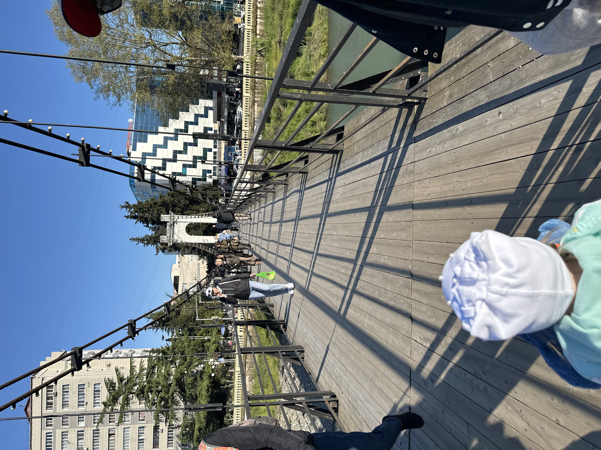 The image captures a sunny day on a pedestrian suspension bridge, offering a view of an urban environment, likely in Sochi, Russia. The bridge features a light-colored wooden deck with black metal railings on both sides, which cast long, distinct shadows, suggesting either morning or late afternoon. Suspension cables with small, round lights are visible running along the length of the bridge.
Several people are present on the bridge. In the mid-ground, a person wearing a black jacket, light blue jeans, and a white baseball cap is walking towards the viewer, holding a bright green plastic bag. Closer to the foreground on the right, a child in a light blue jacket and a white, possibly ruffled or patterned, hat is partially visible. Other pedestrians can be seen further down the bridge.
The setting includes a calm body of water, likely a river or canal, flowing beneath the bridge, with green banks visible. The background features various buildings and abundant greenery. On the left side, a prominent building with a distinctive white and black zig-zag pattern on its facade stands out, with other modern structures nearby. On the right, a more traditional light-colored building with numerous windows is visible. The sky is clear and blue. There is no clearly discernible text on any of the visible signs or buildings due to distance and resolution.