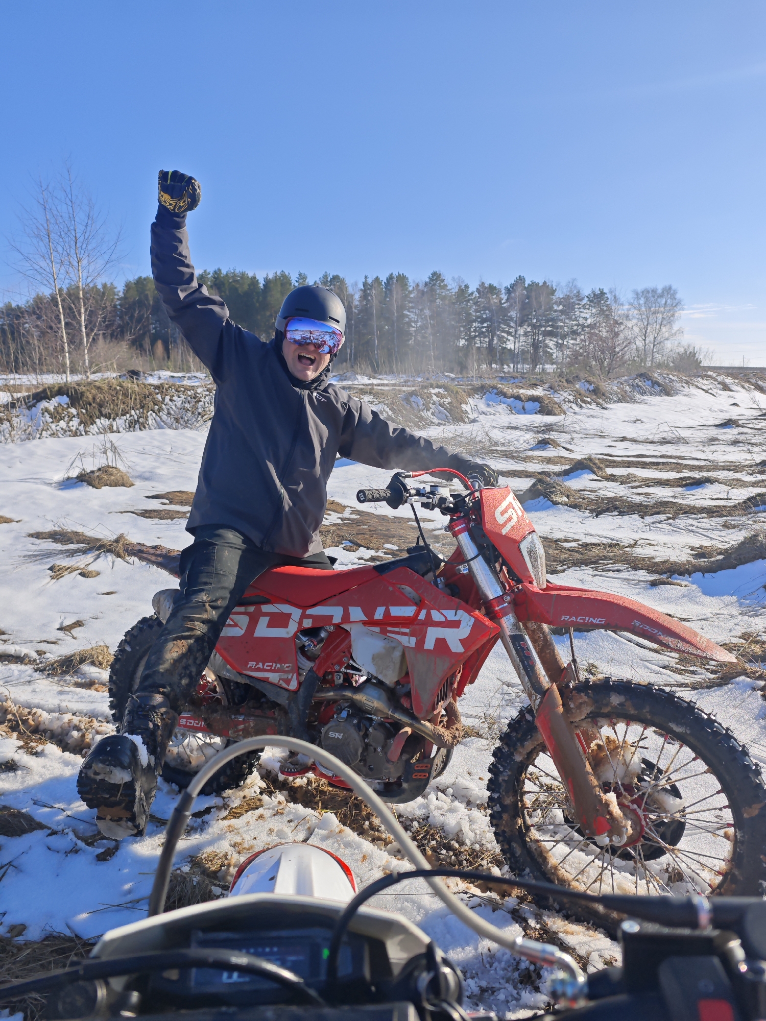 A man is sitting on a red dirt bike in a snowy, rural environment. The man is wearing a black helmet, goggles, and a dark jacket. His left arm is raised in the air in a celebratory gesture. The dirt bike is heavily covered in mud. The background shows a field covered in snow with patches of dry vegetation and a line of trees in the distance under a clear blue sky. In the foreground, parts of another motorcycle are visible. Based on the metadata, the location is Прикамский, Russia.