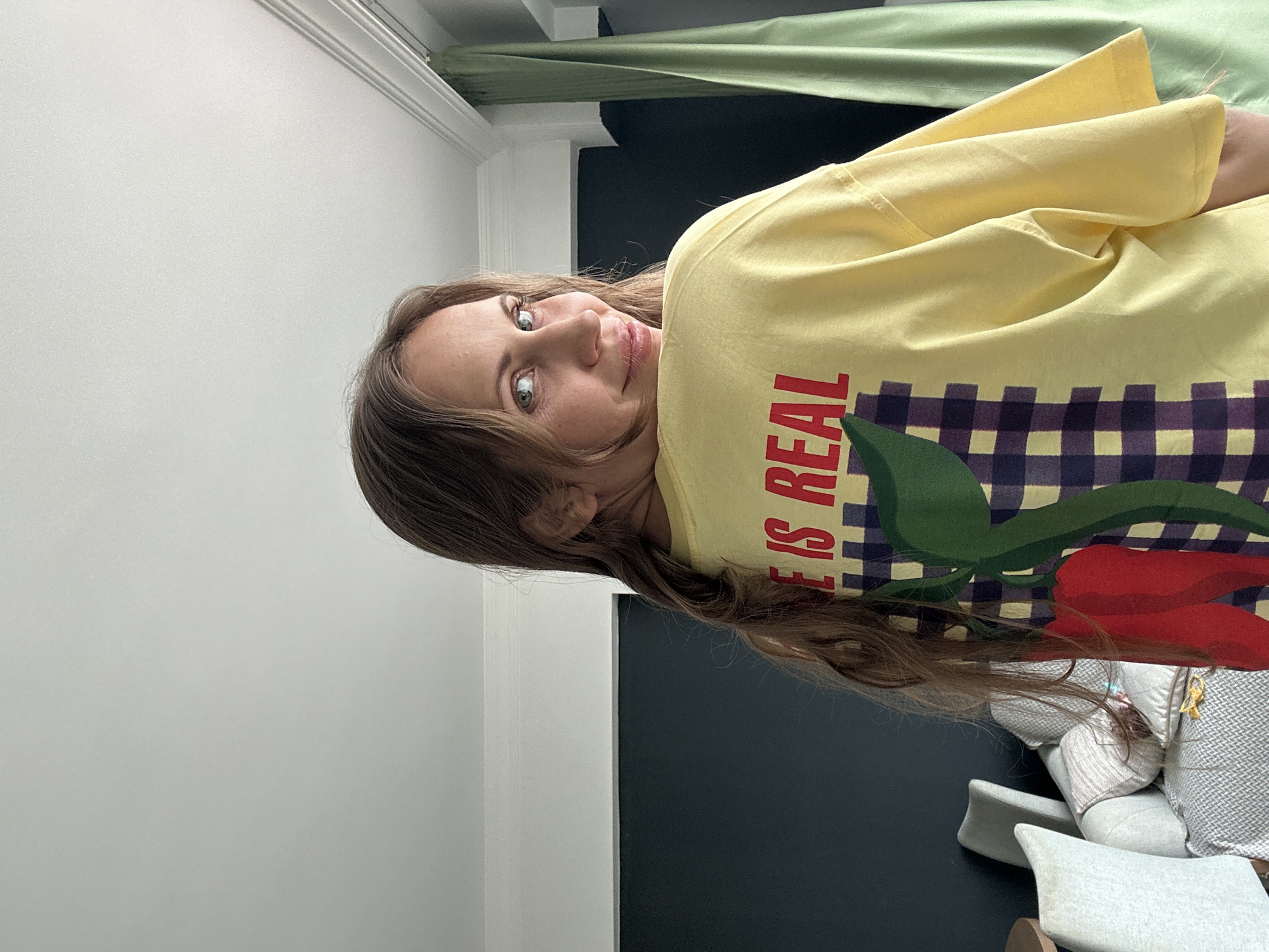 The image shows a woman with light brown hair and blue eyes looking at the camera. She is wearing a yellow t-shirt with the words "LIFE IS REAL" in red letters. A design of chili peppers with a purple and white checkered background is printed on the shirt as well. She is indoors, standing against a white wall. In the background, a dark blue surface can be seen to the right. A light green curtain is visible in the upper portion of the image. Cushions of a greyish color are also visible in the lower right corner. The location in the city of Fethiye, Turkey cannot be determined from this image.