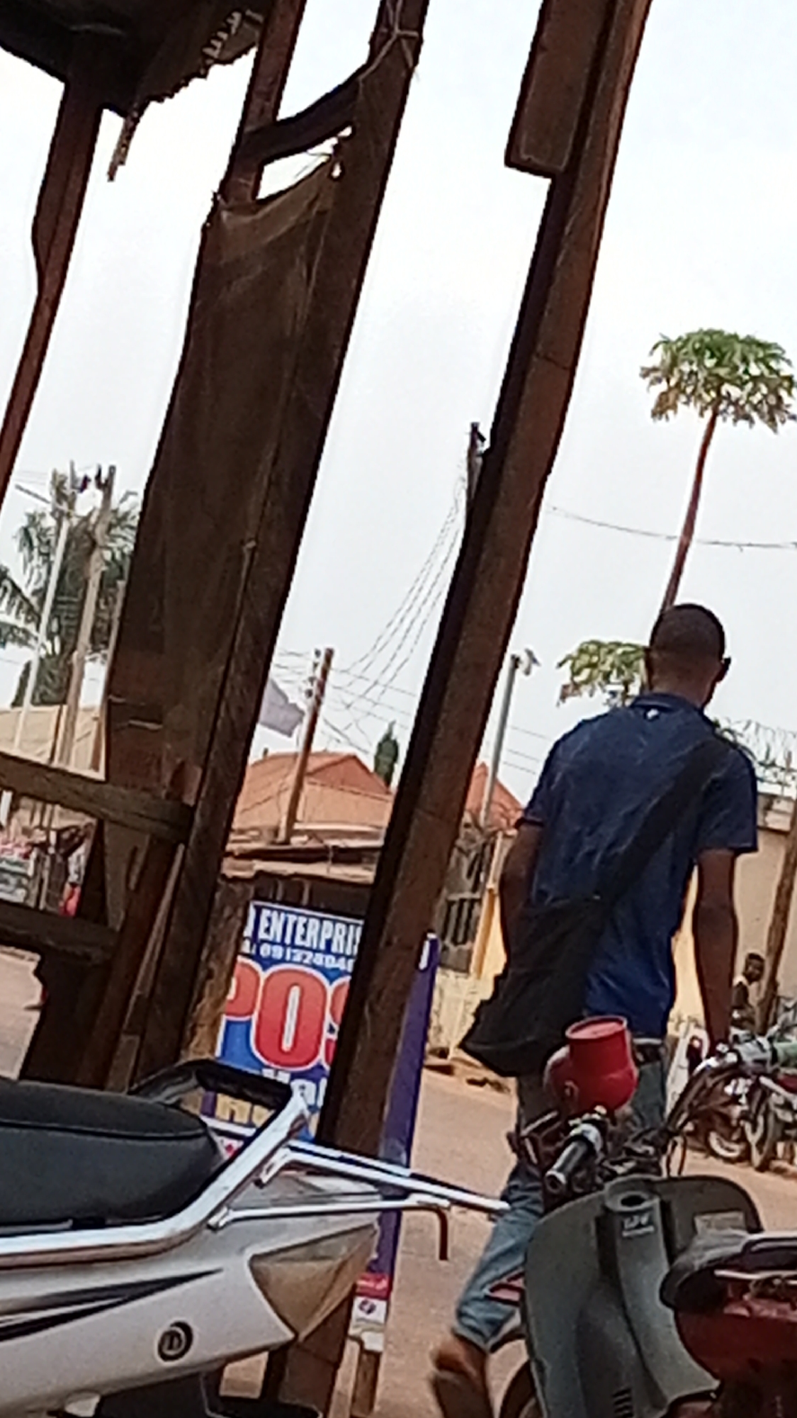 The scene is set in Sabon Tasha, Nigeria, featuring a street view partially obstructed by a wooden structure, likely part of a market stall or roadside shop. The sky is overcast.
A man wearing a blue t-shirt and a black shoulder bag is walking away from the viewer. He is captured from the back.
Several motorcycles are visible, some parked and others partially within the frame. A POS (Point of Sale) advertising sign is partly visible next to the man, displaying "ENTERPRISE" and a phone number. The signage suggests financial services are available.
In the background, there are buildings with corrugated iron roofs and a tall, slender tree with a cluster of leaves at the top. Utility poles and wires are also present.
The immediate foreground contains a white motorcycle, partially obscuring the lower portion of the scene. The scene conveys a sense of everyday life in an urban environment.