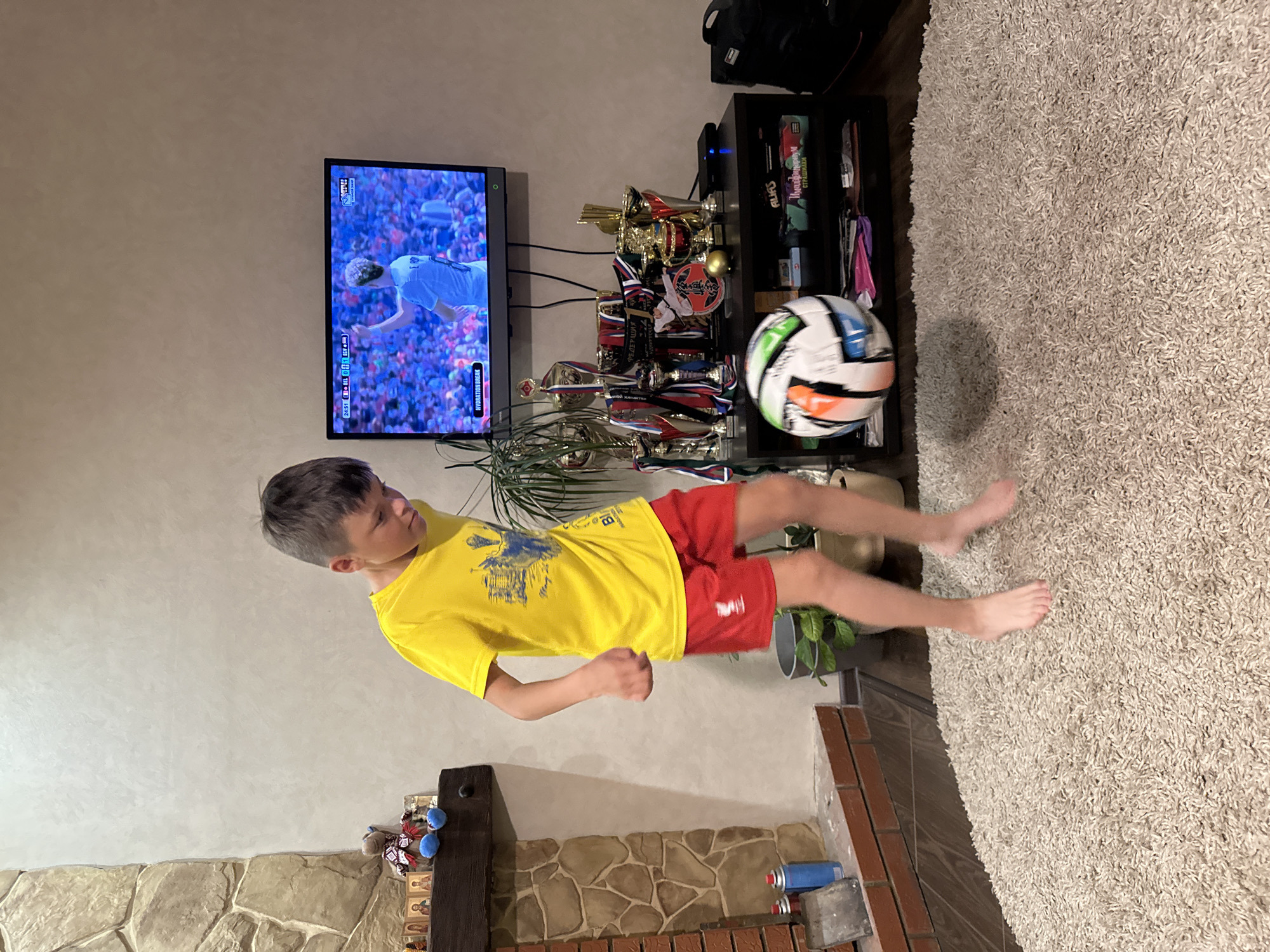 A young boy, wearing a yellow t-shirt and red shorts, is captured mid-action as he juggles a soccer ball on a fluffy beige rug in a living room. The boy's bare feet are on the rug, and his body is angled as he focuses on the ball, which is in the air before him. The room is furnished with a television displaying a soccer match, a black entertainment stand filled with trophies and what appears to be board games, a fireplace with stone detailing, and a potted plant. The overall impression is of a playful, domestic scene, likely in Chaikovsky, Russia, given the context.