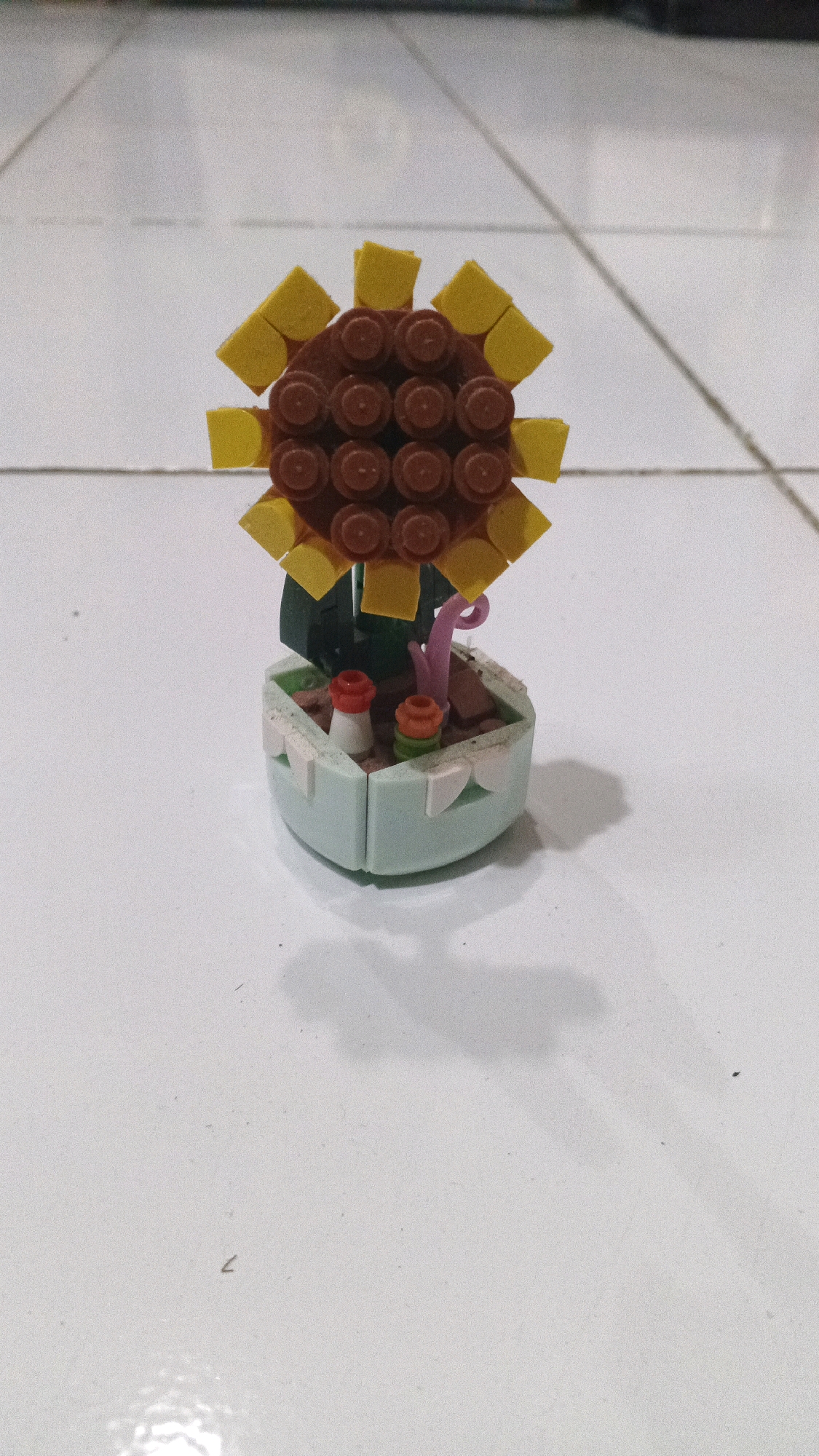 A miniature Lego sunflower is positioned on a white tiled floor. The sunflower features yellow petals surrounding a brown, circular center composed of smaller brown Lego pieces. The stem is green, and it sits in a light blue Lego pot. Inside the pot, two small red and orange Lego pieces are visible, resembling small plants or decorative elements. A pink Lego piece, possibly another stem or decorative element, is also present in the pot. The floor tiles show grout lines, and a shadow of the Lego sunflower is cast on the surface. The overall scene is a close-up, head-on view of the toy. The specific location in Winong, Indonesia, is not discernible from the image itself.