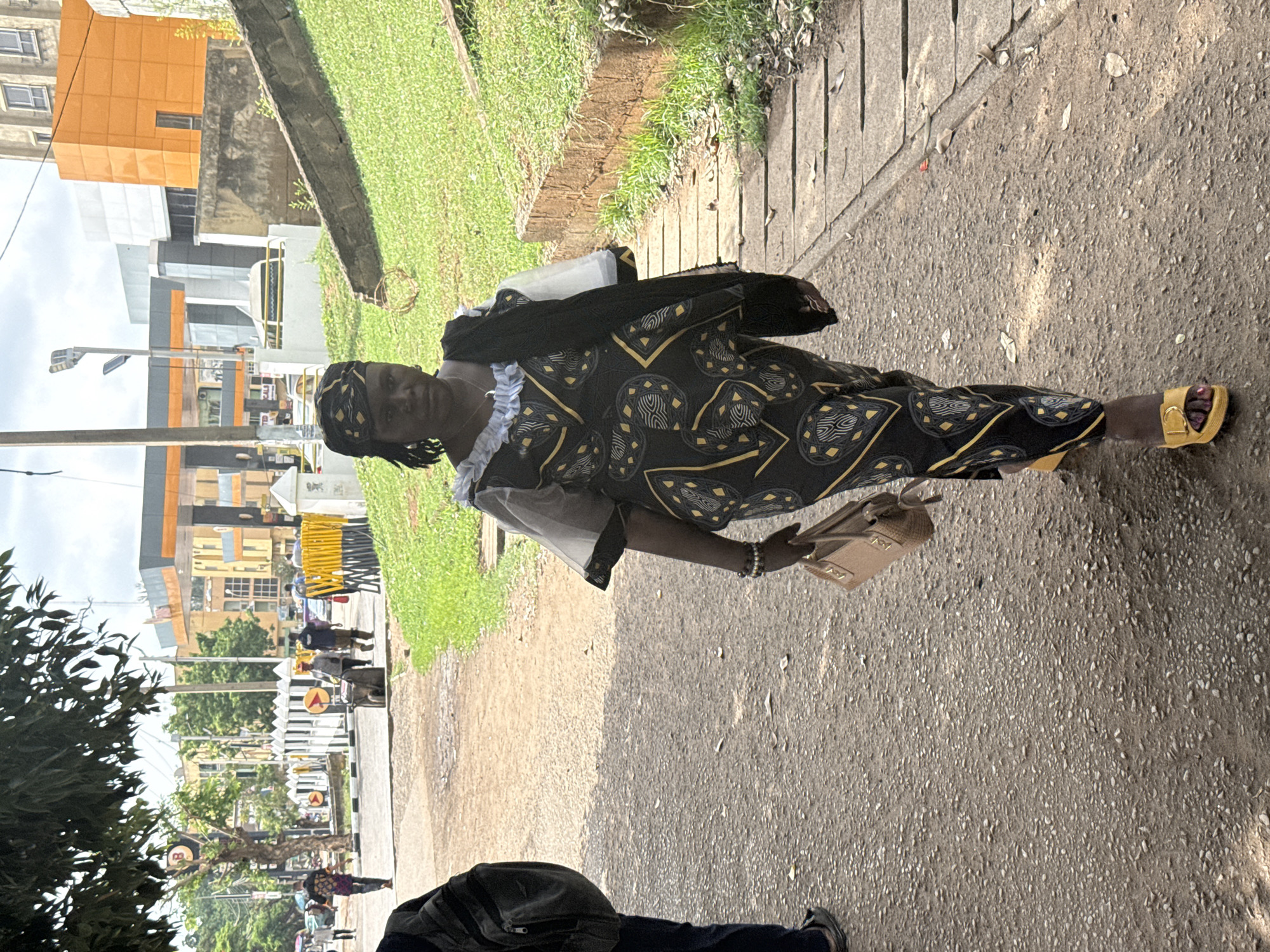 A woman stands on a paved walkway near a street in Abuja, Nigeria. She is wearing a patterned black and gold dress, yellow sandals, and carrying a tan handbag. A black scarf drapes over her shoulders, and she has a headwrap. Behind her, there is grass, and buildings with orange and gray facades are visible across the street. There are other pedestrians in the background. The walkway is adjacent to a curb and street. Another person is partially visible at the bottom of the frame.