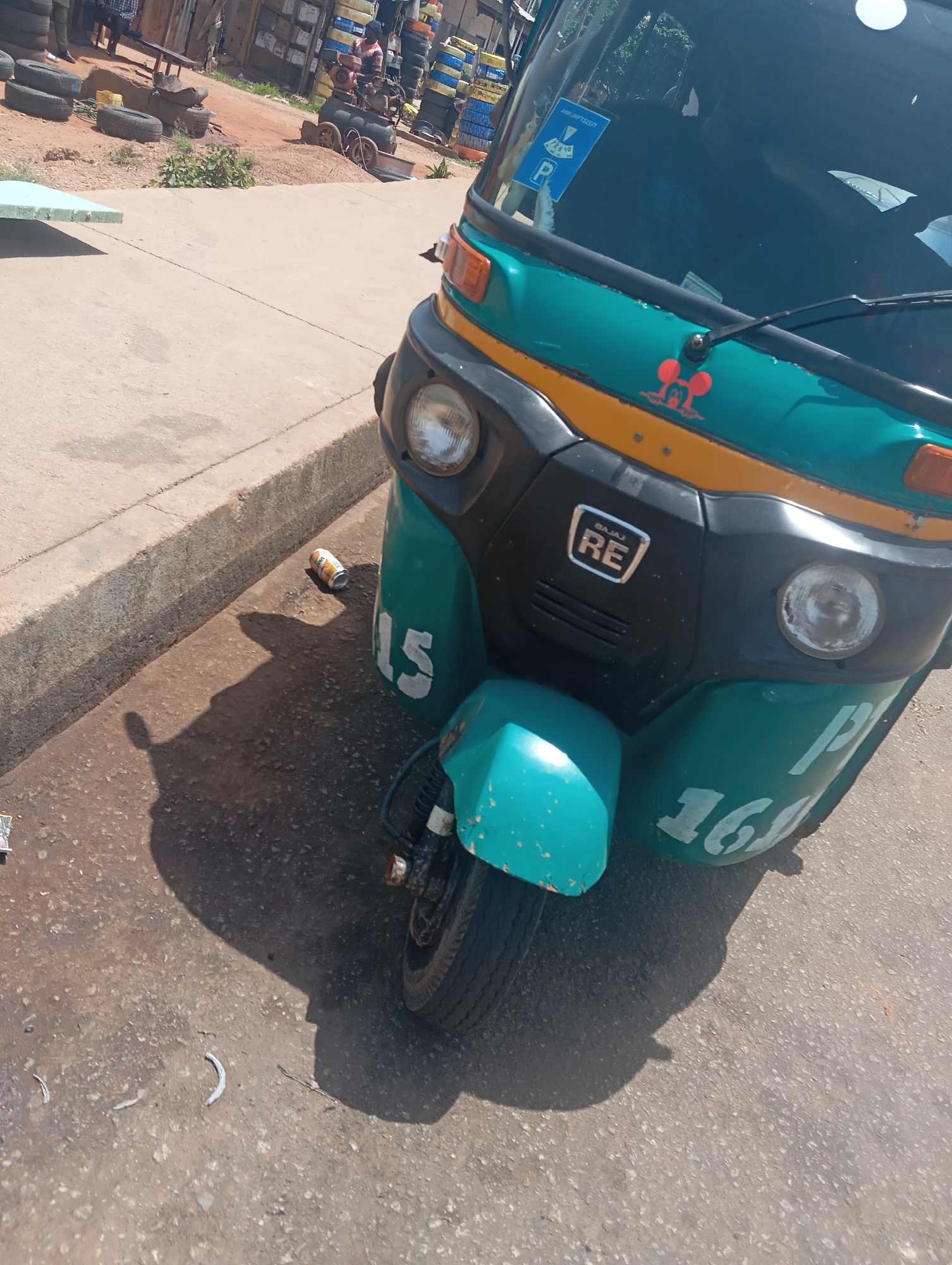 The image shows an outdoor scene in the city of Jos, Nigeria, with a teal and orange auto-rickshaw as the primary subject. The auto-rickshaw is parked on a paved surface next to a curb. It has a large headlight, a windshield with a parking permit sticker, and markings on its body that appear to be numbers, including "15" and "161". A discarded beverage can is visible on the ground near the auto-rickshaw. In the background, stacks of tires are piled high, suggesting a tire repair shop or a storage area for used tires. There are also people in the background, partially visible, and some wooden structures. The overall environment appears to be a commercial or industrial area.