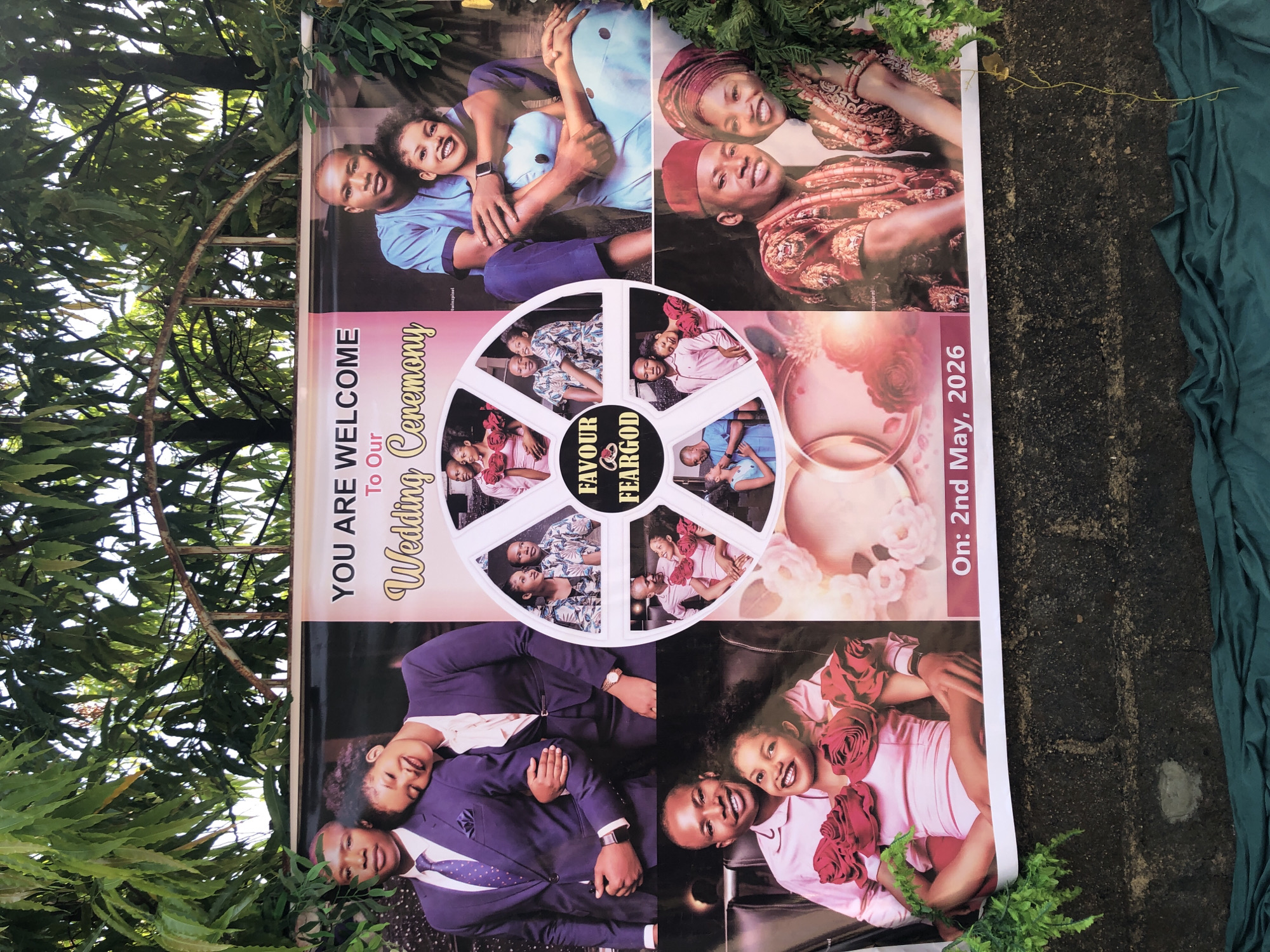 Here is a description of the image:
The image shows a wedding welcome sign, placed outdoors, likely in Akwanga, Nigeria. The sign features a montage of photos of the couple, with the names "Favour" and "Feargod," and the wedding date "On: 2nd May, 2026" printed on it. The text "YOU ARE WELCOME To Our Wedding Ceremony" is prominently displayed. The sign is surrounded by greenery. The background is blurry.
