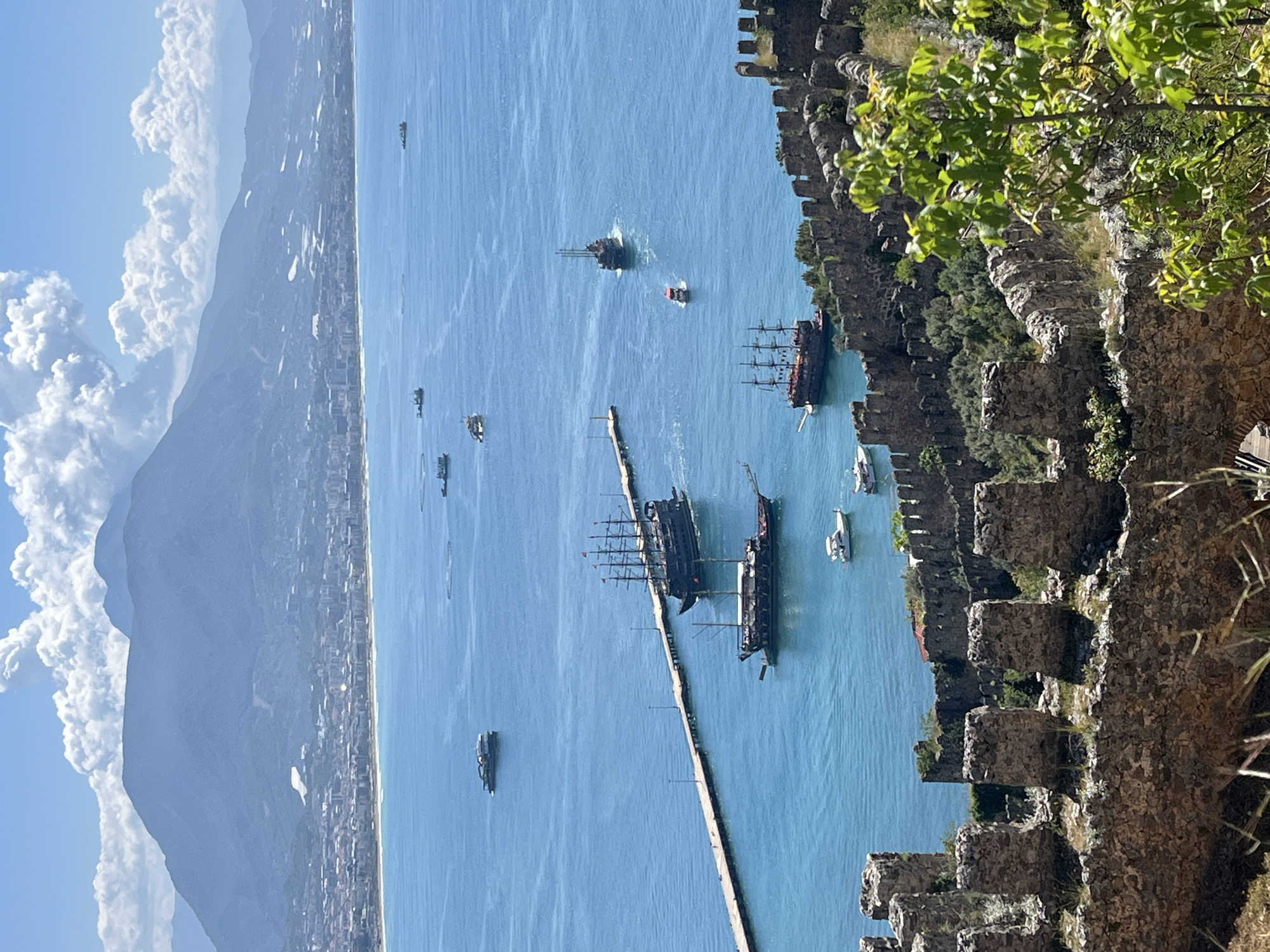 The image captures a view from Alanya Castle in Alanya, Turkey. The foreground features a section of the castle walls, constructed of light brown stone blocks. A bit of foliage hangs from the top right of the image. The middle ground shows the harbor and the sea. There are several boats of various sizes on the water, including a number of pirate-themed ships. A long, narrow pier stretches into the water. The background displays the coastline of Alanya with buildings lining the shore. Mountains are visible further inland, partially obscured by clouds. The sky is blue, with some cumulus clouds.