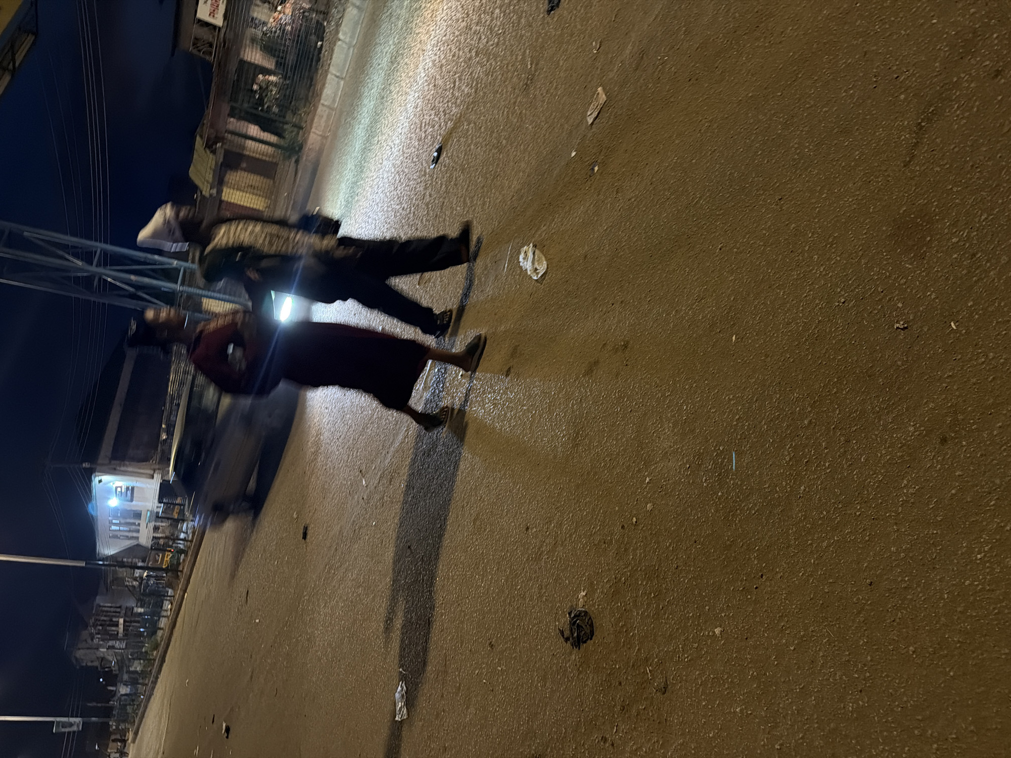 Two individuals are captured in motion on a wet, asphalt road at night. The person on the left, wearing a dark red garment and grey sandals, walks with their body angled towards the right. Beside them, to the right, another individual is seen in profile, wearing dark trousers, a patterned top, and a headwrap. This person also walks towards the right, with their left leg forward and their right arm bent.
The environment suggests an urban setting in Bukuru, Nigeria. The road surface is damp, reflecting the ambient light sources. Scattered debris, including paper and what appears to be a black plastic bag, litters the road. In the background, blurred by motion and low light, are the outlines of buildings with illuminated windows. A car, its headlights on, is also visible moving away from the camera, creating a streak of light on the wet road. Overhead, a metal structure, possibly part of an overhead road sign or infrastructure, is visible against the dark night sky. The lighting is predominantly artificial, casting long shadows and creating a somewhat grainy image quality due to the low light conditions.