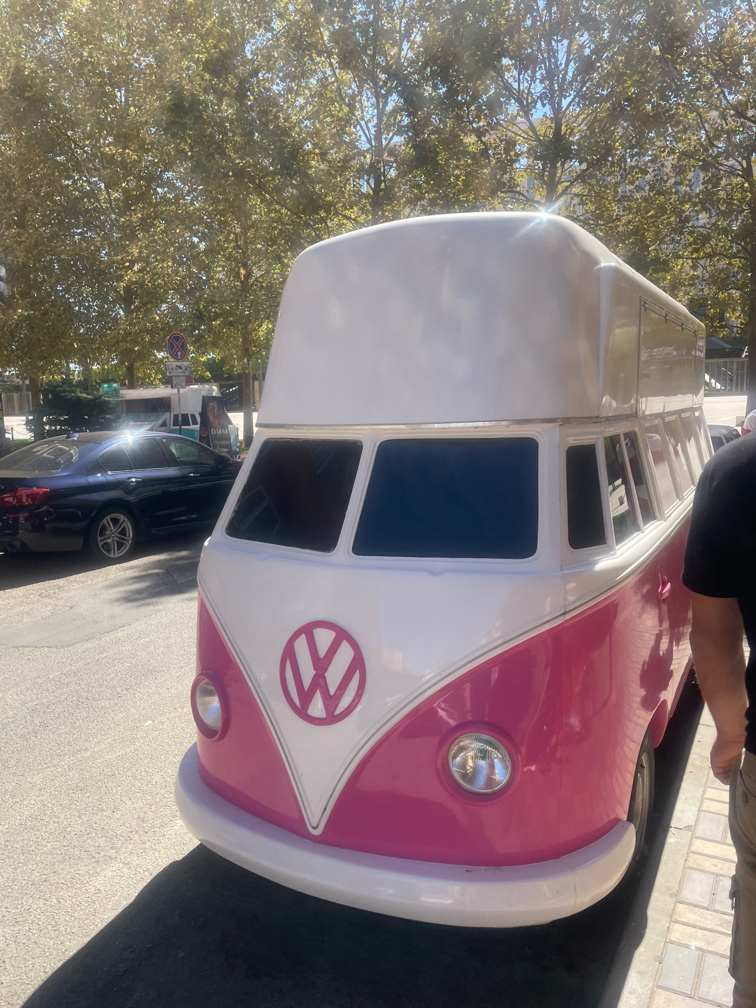 A vintage Volkswagen bus, painted pink and white, is parked on a city street in Krasnodar, Russia. The bus has a distinctive rounded shape, with a prominent white raised roof. The front of the bus features a pink V-shaped emblem with the Volkswagen logo. In the background, a dark blue sedan is parked, and trees with green and yellow leaves line the street. A person, seen from the waist down, stands next to the bus. The scene is illuminated by sunlight, casting shadows on the pavement.
