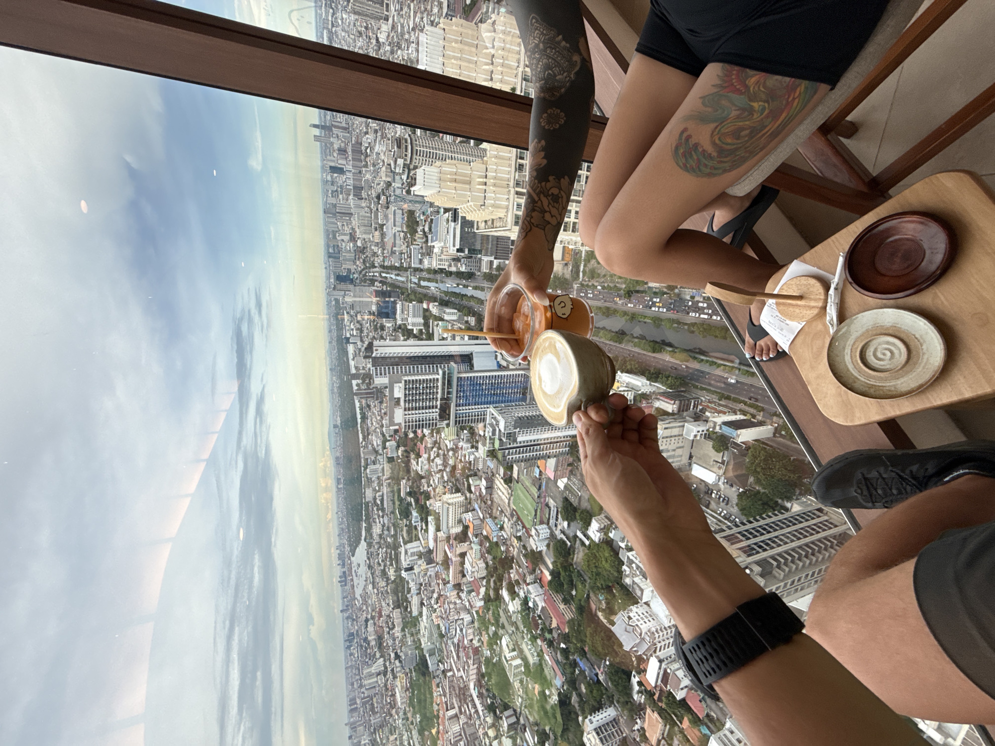 The image shows a view from a high-rise location in Bangkok, Thailand. Two people are positioned at a table by a large window overlooking the city. One person is holding a cup of coffee, while the other is holding a plastic cup with a straw and orange liquid. Both individuals have tattoos on their arms and legs. On the table are small plates and a wooden tray, along with a receipt. The cityscape below features a mix of buildings, roads, and waterways, stretching out to the horizon where the sky is visible through a glass window.