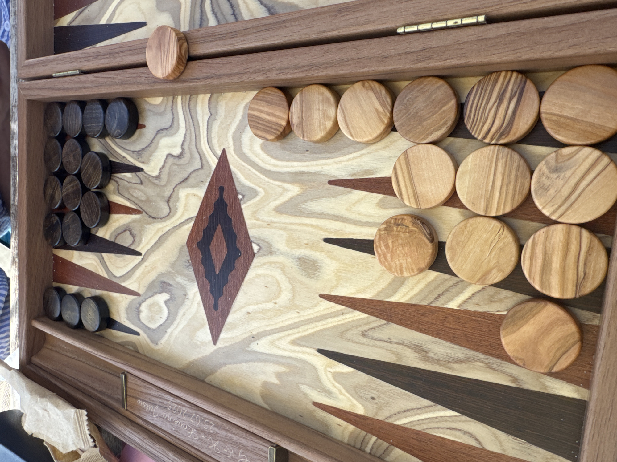The image shows a close-up view of a wooden backgammon board. The board has a light-colored wooden surface with dark brown and reddish-brown triangular markings. There are two sets of playing pieces, or checkers. One set consists of dark brown, circular checkers arranged in stacks on the upper left side of the board. The other set consists of light brown, circular checkers arranged in stacks on the right side of the board. One of the light brown checkers is positioned on the edge of the board. There is also a wooden strip on the left side of the board with some engraved text, which appears to be a name and a date: "S-K-Leon no Futsu 25.07.2025". The background is out of focus, but appears to be a striped fabric. The scene is indoors. There is no information in the image that allows for the location in the city of Pallini Municipal Unit, Greece.
