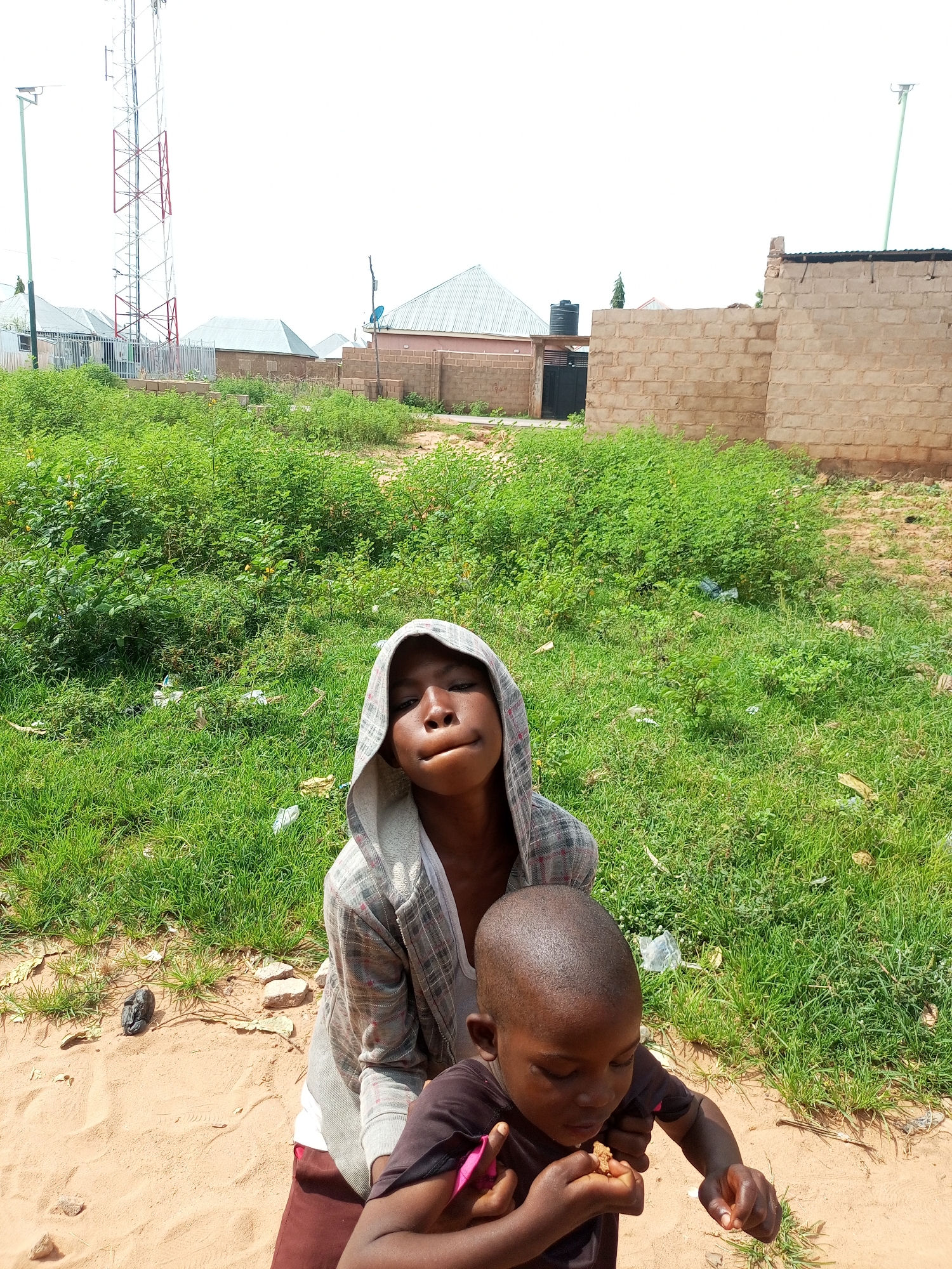 In Tempure, Nigeria, two children are captured in a photograph. The foreground features two young individuals, one older child wearing a hooded gray and white checkered jacket and the other a younger child with a dark t-shirt. The older child is looking directly at the camera with a somewhat quizzical expression, while the younger child is focused on eating a piece of food held in their hands.
The background depicts a semi-urban environment. A tall, metal telecommunications tower stands prominently on the left side of the frame. Surrounding this are several buildings with metallic roofs, typical of residential structures in the area. A brick wall, constructed from light-colored blocks, dominates the right side of the background, with a portion appearing incomplete or under construction.
The ground in the foreground consists of a sandy, earthy surface interspersed with patches of green grass. Some scattered debris, including plastic bags and small stones, is visible on the ground and within the grassy areas. The overall scene suggests a bright, outdoor setting, likely during daylight hours.
