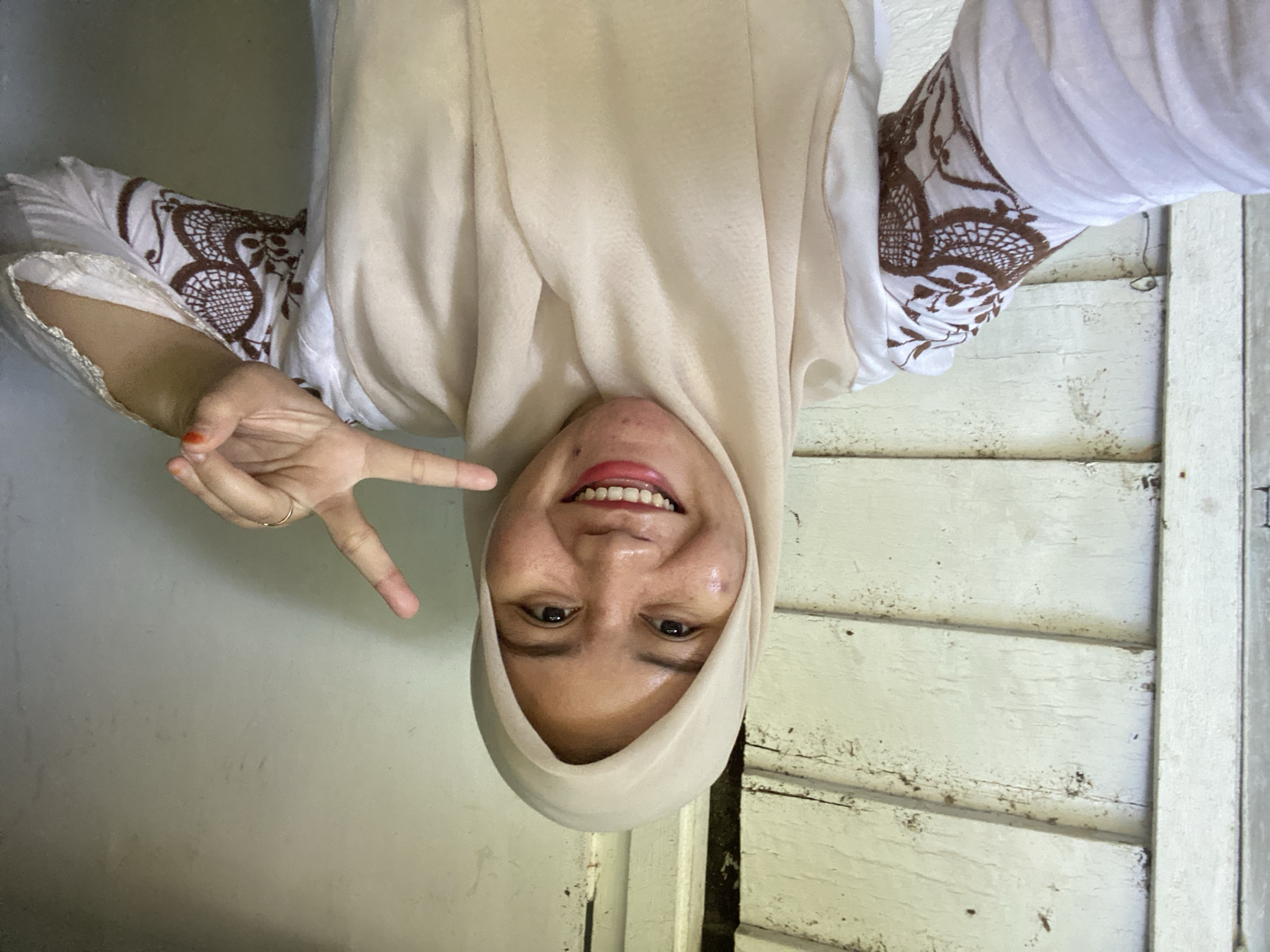 A young woman wearing a light beige hijab and a white shirt with brown lace-like patterns on the sleeves is taking a selfie. She is smiling broadly and holding up two fingers in a peace sign with her right hand. Her left hand is not visible. The background appears to be the exterior of a wooden building, possibly a house, with weathered white horizontal planks. The lighting suggests it is daytime. The image was likely taken in a residential area of Banjarbaru, Indonesia, given the architectural style and common attire.