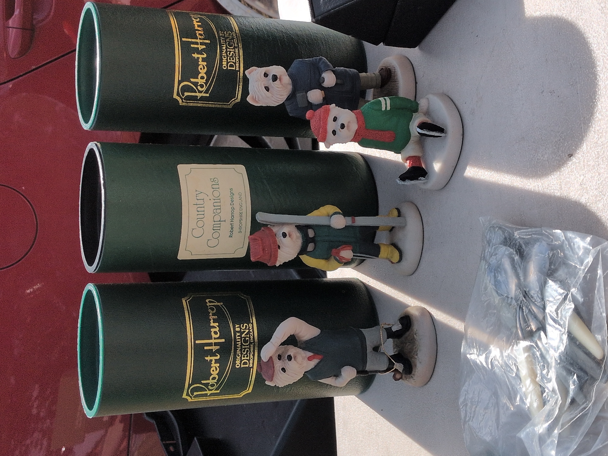 The image shows four decorative figurines of West Highland White Terriers, each dressed in different attire. The figurines are positioned in front of three green cylindrical containers, two of which have gold labels that read "Robert Harrop" and "Originality by Designs Shropshire, England." One of the containers has a label that reads "Country Companions." The figurines include one wearing a hat, vest, and tie, holding a golf club; another dressed in winter clothing with skis; a third wearing a red hat and scarf with a green sweater; and the fourth standing in a dark blue jacket and brown pants, holding two objects that appear to be tools. In the foreground, a clear plastic bag contains what appear to be silver utensils. The items are displayed on a light-colored, textured surface, possibly a table, with strong shadows cast by the objects and sunlight. The background features a blurry, dark red surface, likely a vehicle. The location is specified as Claverley, United Kingdom.