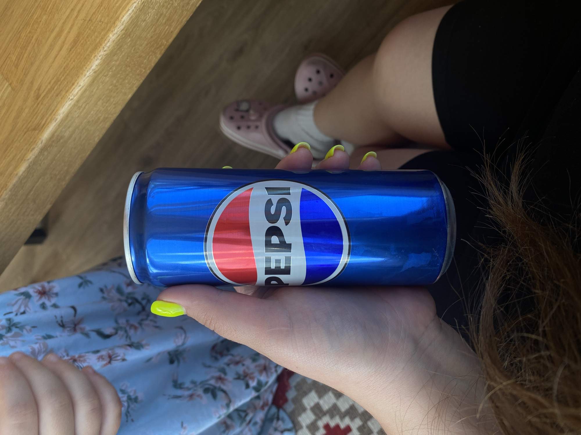 A person's hand, with bright neon yellow fingernails, holds a metallic blue Pepsi can. The can features the iconic Pepsi logo, a red, white, and blue circle with the word "PEPSI" in white text. In the background, a person's legs are visible, wearing pink Crocs and white socks. They are sitting on a wooden surface, possibly a table or bench, with a patterned rug beneath them. A portion of a floral blue dress is also visible. The scene appears to be indoors, in what could be a casual setting. No specific location in Peterhof, Russia, is discernible from the image.