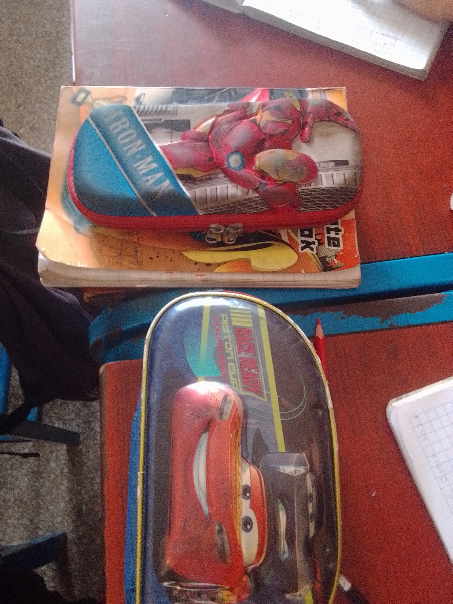 A low-angle, slightly blurry medium shot shows a stack of items on a red wooden surface. The top item is a pencil case featuring Iron Man. It is a rectangular case with a red zipper and a blue flap that displays "IRON-MAN" in white text. The front of the pencil case has a detailed image of Iron Man in a red and gold suit. This pencil case rests on top of a notebook with orange and black graphics. Below this is another pencil case, this one featuring Lightning McQueen from the movie "Cars." This case is also rectangular, with a clear plastic front revealing the character. Lightning McQueen is depicted in a race car design with the text "RACE READY PISTON CUP" visible. A red pencil lies to the right of this second pencil case. The background shows a textured, greyish wall and a glimpse of blue furniture. The scene appears to be an indoor setting, possibly a desk in a home or classroom. No specific location in La Tagua, Venezuela, can be identified from the visual information provided.