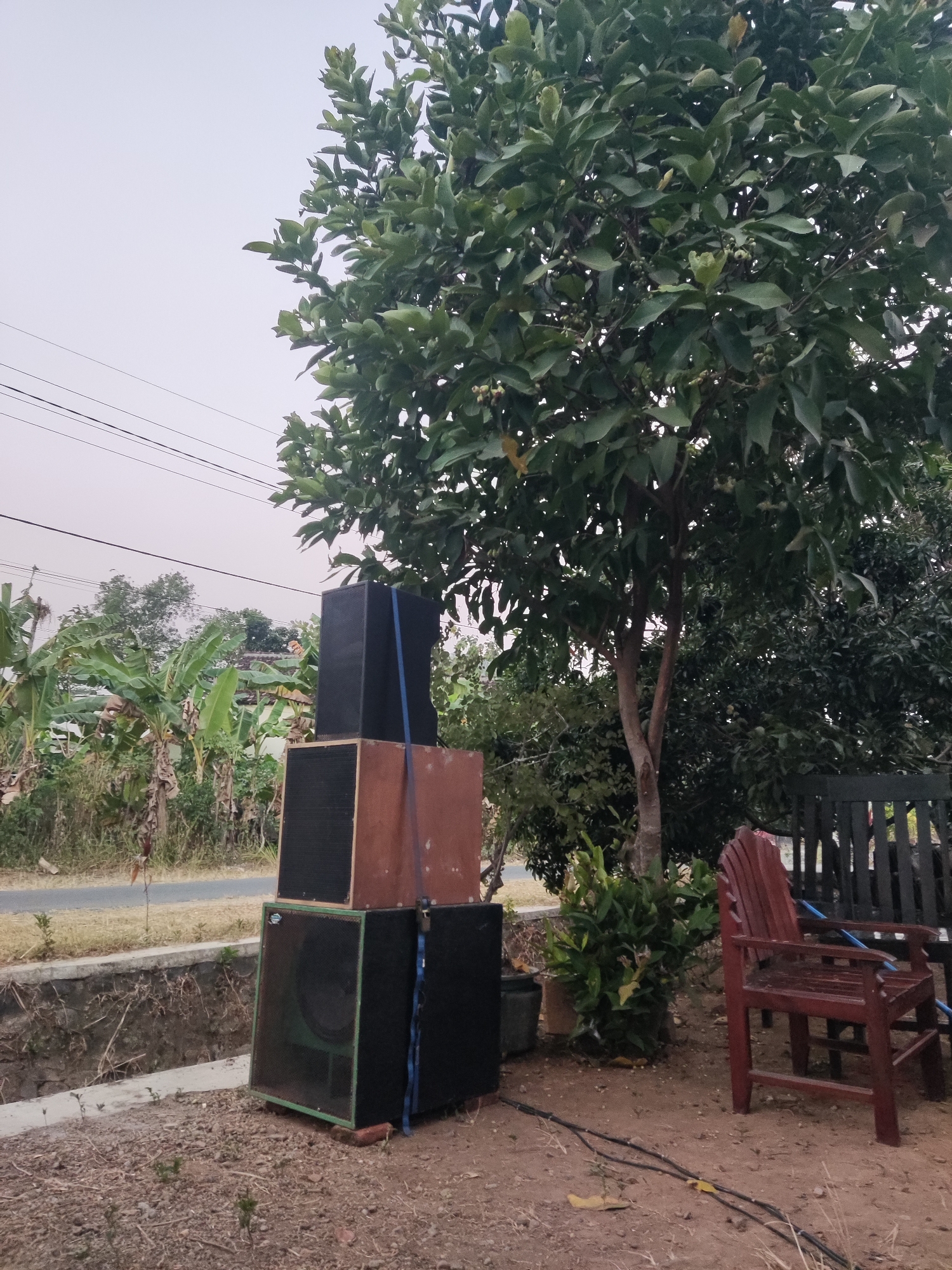 In Ngentrong, Indonesia, the image depicts an outdoor scene with a stack of four speakers in the foreground. The speakers are arranged vertically, with the bottom speaker being the largest and having a green grille. Above it sits a speaker with a brown wooden enclosure and a black grille, followed by another speaker with a black grille and a blue strap. The top speaker is entirely black. To the right of the speakers, there is a red wooden chair and a black wooden bench. Behind the speakers, a large tree with dense green foliage dominates the right side of the frame. In the background, there are banana trees, other vegetation, a road, and power lines stretching across the sky. The ground is covered with dry leaves and dirt.