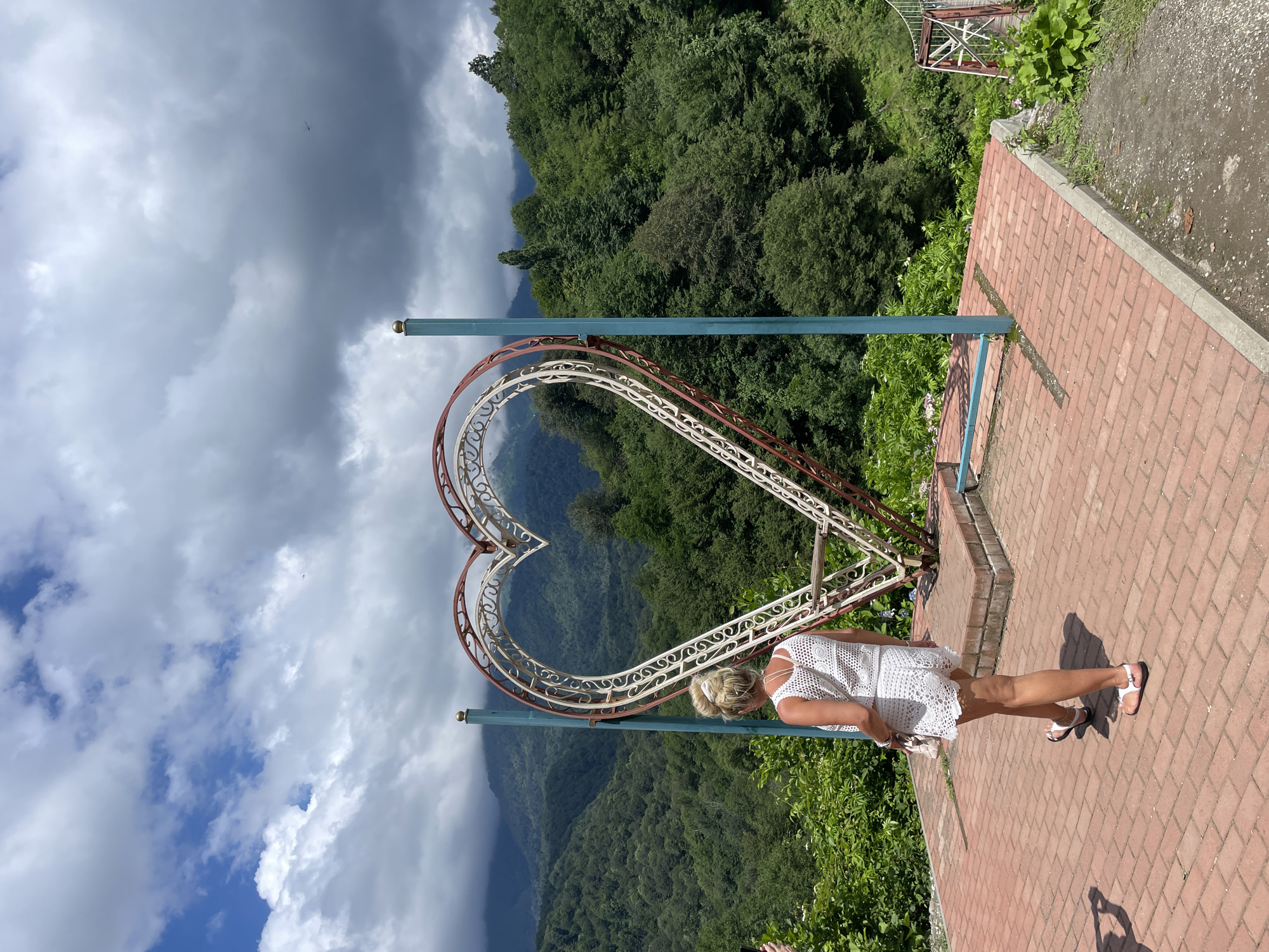 A woman with blonde hair pulled back in a bun, wearing a white crochet top and shorts, walks away from the camera on a red brick pathway. She is positioned to the left of a large, decorative metal heart structure. The heart is framed by two teal posts and features intricate white and red metal scrollwork. Behind the heart, a lush, green, mountainous landscape unfolds under a cloudy sky. The mountains are densely covered with trees. To the right of the scene, a portion of a suspension bridge is visible. The woman is carrying a light-colored purse on her left shoulder. The pathway is bordered by greenery and a dirt path.