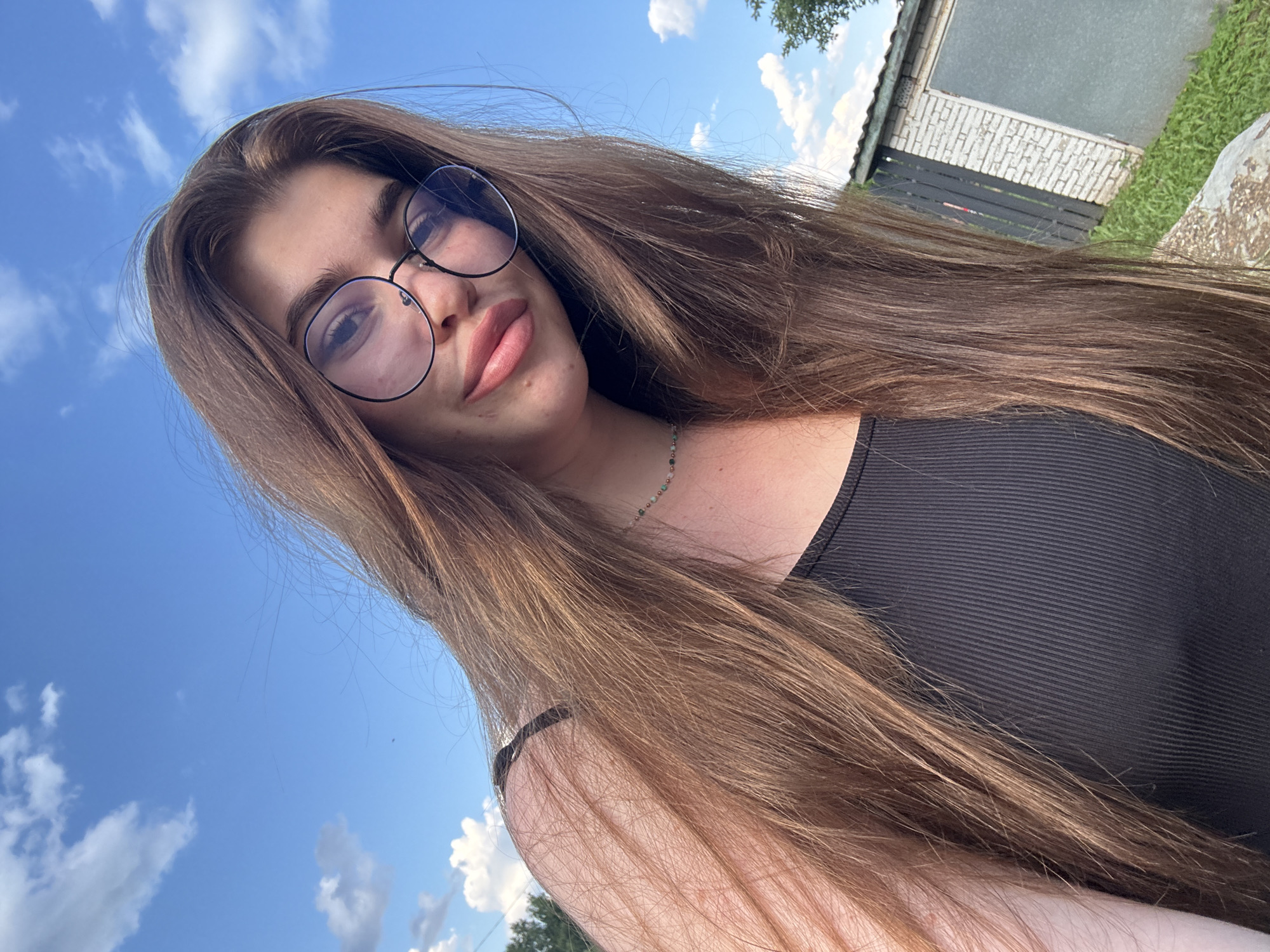 A young woman with long, brown hair and glasses is taking a selfie outdoors. She is wearing a black tank top and a necklace. The background shows a blue sky with clouds, trees, and a building with a fence. The lighting suggests it is daytime. The overall impression is a casual, informal portrait. There are no discernible landmarks that would place this image specifically in the city of Мещёры, Russia.