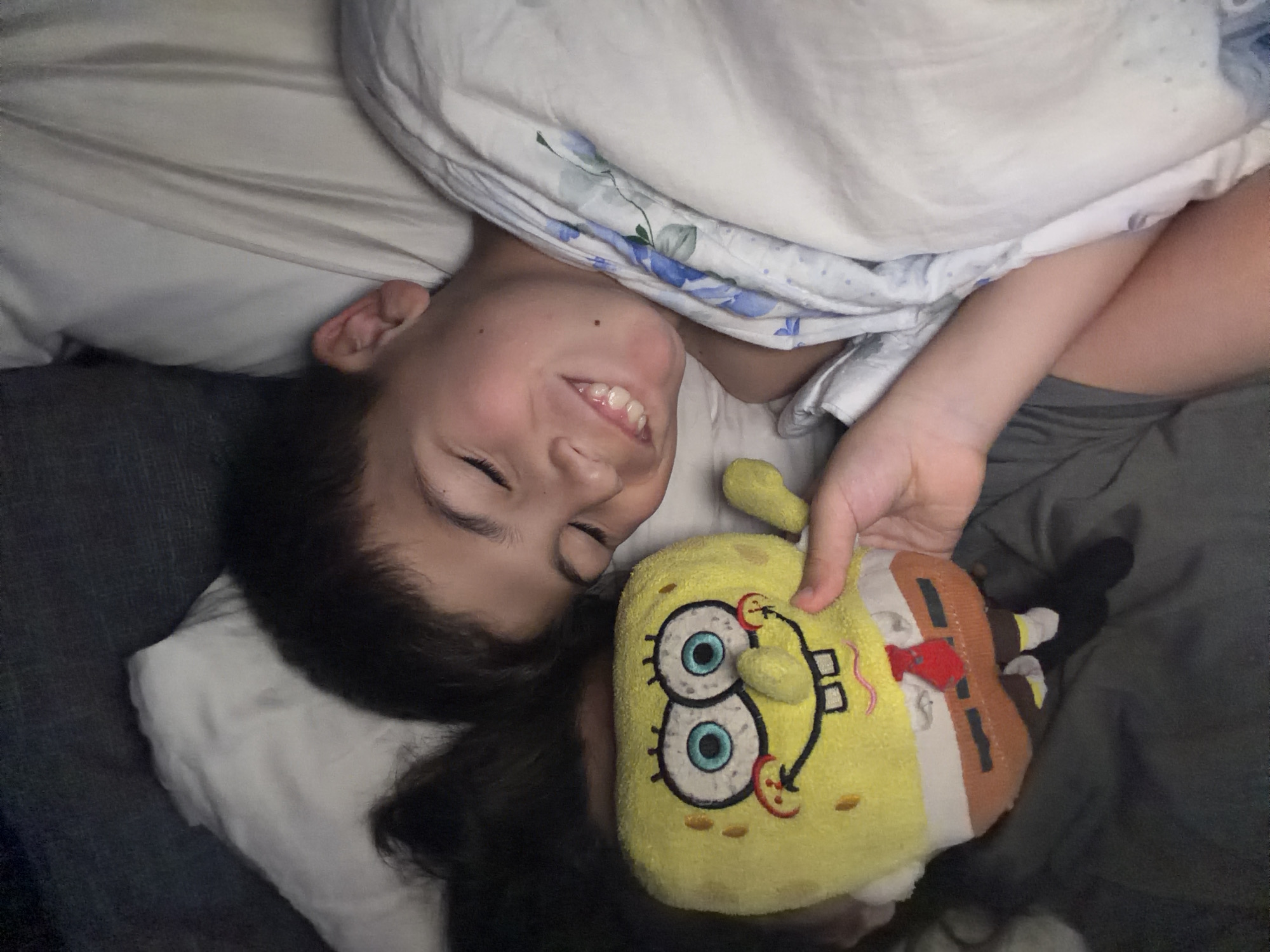 A young boy lies in a bed with white sheets and a blanket with blue floral patterns. He is positioned on his left side, smiling with his eyes open, and holding a Spongebob Squarepants plush toy. The boy has short, dark hair. The bed appears to be indoors. There are no indicators relating to Budva, Montenegro in the scene.