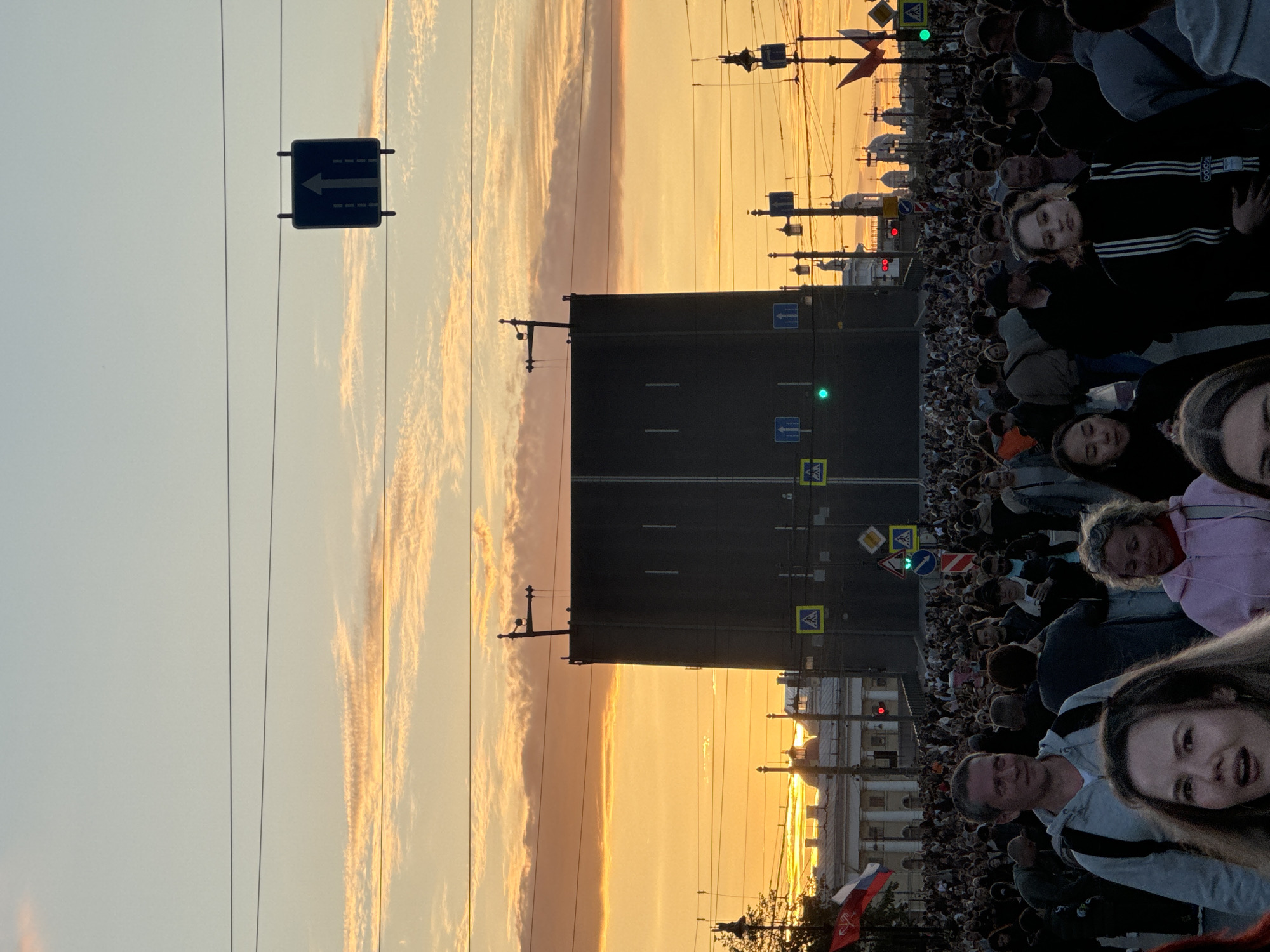 In Saint Petersburg, Russia, at sunset, a crowd of people are gathered on a bridge. The sky is filled with orange and yellow clouds. A large, dark bridge structure dominates the center of the image. Traffic lights and street signs are visible. Several people are in the foreground, looking towards the camera. The atmosphere appears to be festive or celebratory.