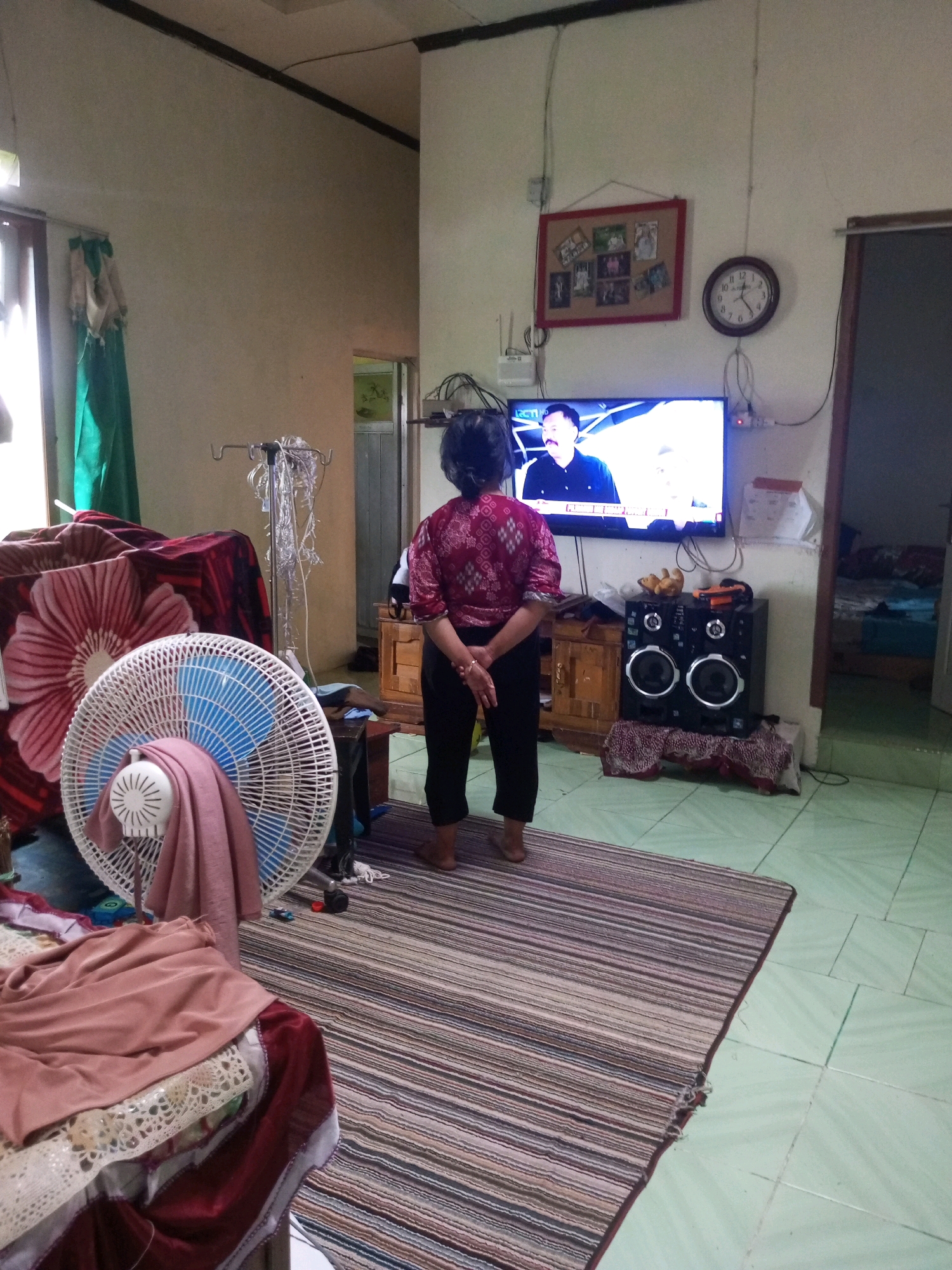 A woman wearing a patterned maroon shirt and black pants stands facing a television in a room. The television displays a news program with a man in a black suit. To the left of the television is a wooden cabinet. To the right of the television is a large black speaker system with two circular speakers. A round clock with Roman numerals hangs on the wall above the television. In front of the woman is a striped rug. To the left of the rug is a white electric fan with a pink cloth draped over it. There is a large red blanket with a floral pattern on a surface behind the fan. The room has light-colored walls and a light-colored tiled floor. A doorway is visible in the background. The scene appears to be an indoor living space in Krasak, Indonesia.