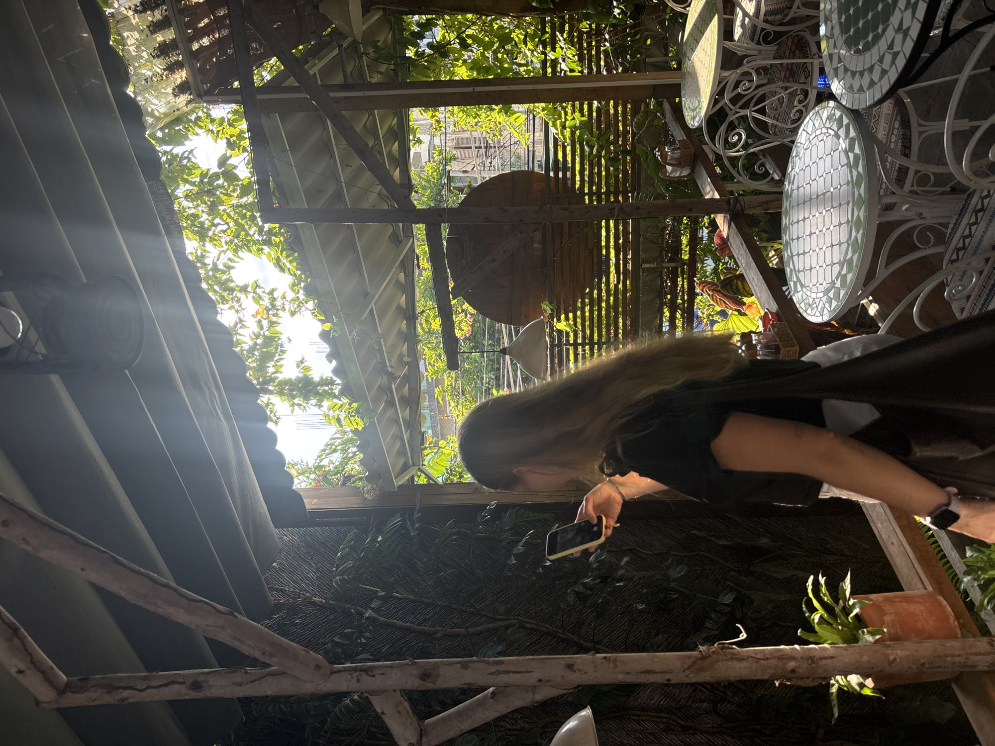 A woman with long blonde hair, wearing a black t-shirt and white pants, stands on a wooden balcony, holding a yellow smartphone in her right hand. She is looking at the phone and appears to be taking a photo or video. The balcony is covered by a slatted roof with sunlight filtering through, illuminating the scene. Lush green foliage surrounds the balcony, with plants in terracotta pots visible.
In the background, there are several outdoor tables and chairs. The tables have white wrought-iron bases and circular mosaic tops with green and white geometric patterns. The chairs also have white wrought-iron frames and patterned cushions. Beyond the balcony, buildings can be seen, suggesting an urban environment.
The overall impression is of a casual outdoor seating area, possibly a cafe or restaurant, in a tropical or subtropical location. The presence of plants and the open-air design suggest a pleasant and relaxed atmosphere. The sunlight creates strong highlights and shadows, adding depth and visual interest to the image. The location is identified as Nha Trang Ward, Vietnam.