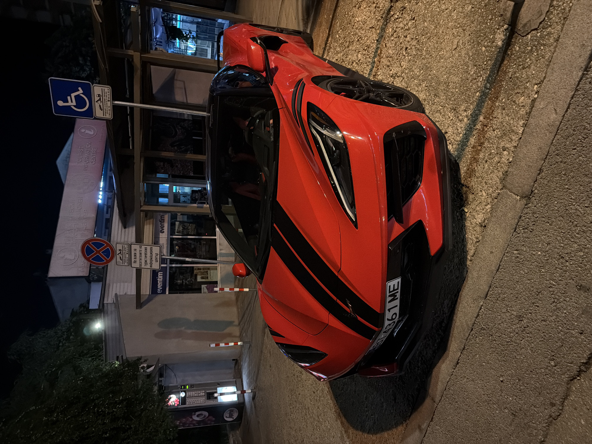 In Veliko Tarnovo, Bulgaria, at night, a vibrant red sports car, a Chevrolet Corvette C8, is parked on a gravel surface next to a paved sidewalk. The car features black racing stripes on its hood and roof. In the background, a building with glass-fronted entrances and prominent "eventim" signage is visible. A white and blue sign indicating a disabled parking spot is mounted on a pole. Another sign with a red circle and a horizontal bar signifies a no-parking zone. There are no people visible in the image. The scene is illuminated by artificial lighting from the building and streetlights.