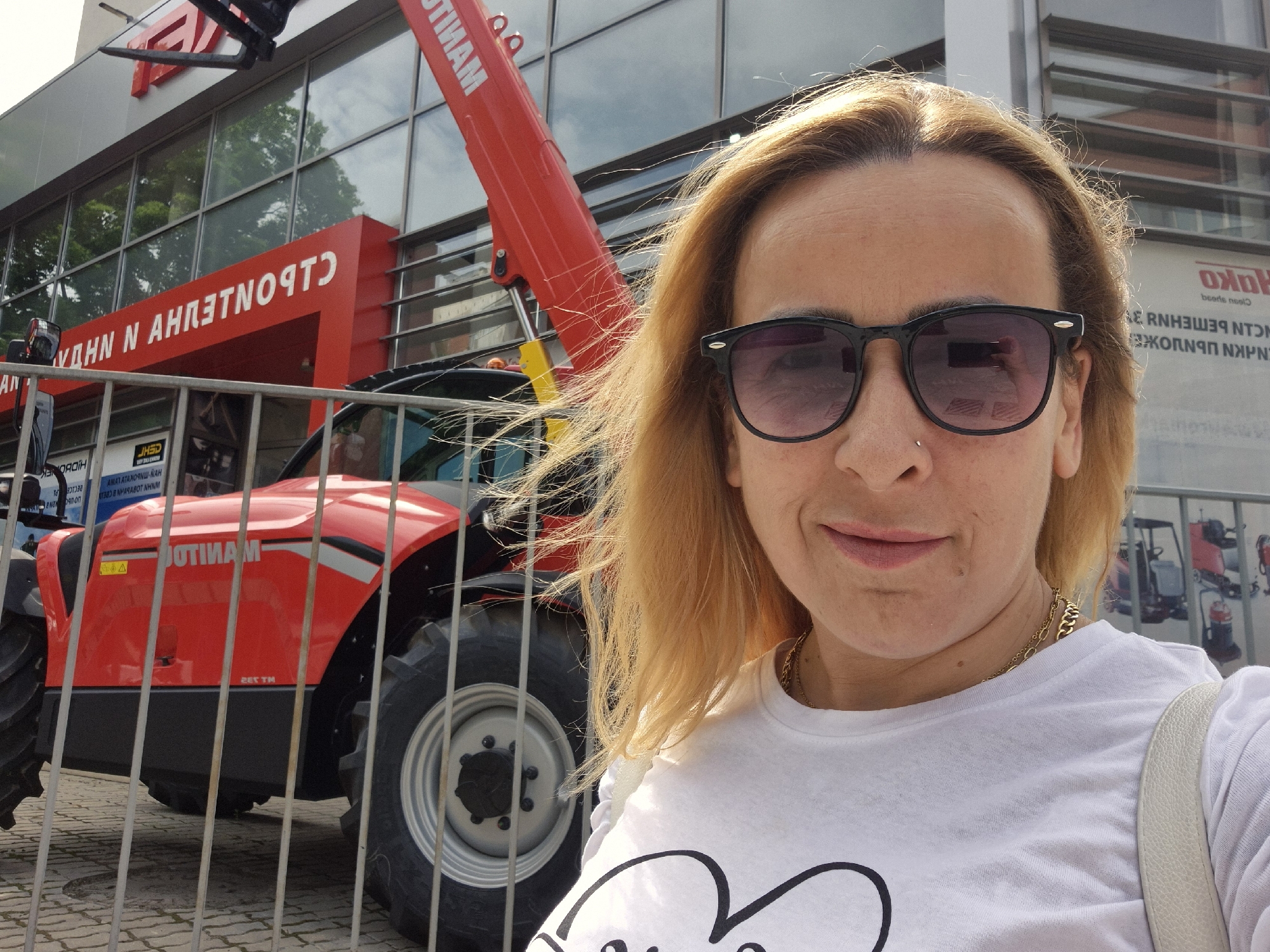 The image is a selfie of a woman wearing sunglasses and a white t-shirt with a design on it. She has blonde hair. In the background, there's a red "MANITOU" construction vehicle, partially visible behind a metal fence. The setting appears to be outdoors in a city, as evidenced by the reflections in the building's windows and signage written in a Cyrillic alphabet. The building's sign above red construction vehicle reads "СТРОИТЕЛНИ МАШИНИ". There is other signage that reads "HAKO" and "beerts neel"