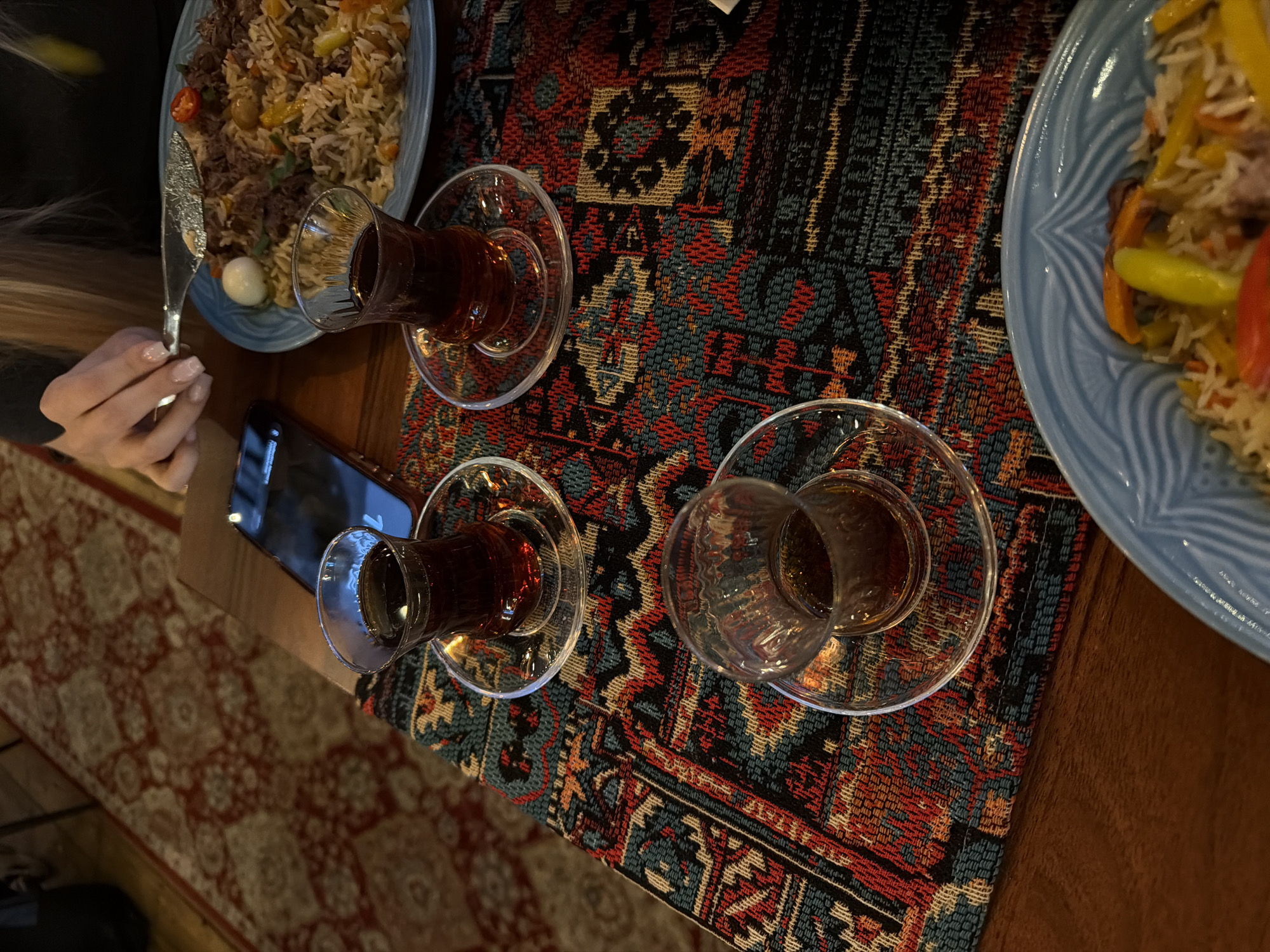The image depicts a table setting, likely in a restaurant, in Warsaw, Poland. The table is covered with a patterned runner featuring geometric designs in red, blue, and black. Three armudu glasses, each filled with tea and resting on glass saucers, are arranged on the runner. A smartphone is placed near one of the glasses. Two blue-rimmed plates containing pilaf are visible on either side of the runner. A person's hand holding a knife is seen near the plate on the left. The flooring below the table is carpeted with a red and beige pattern. The table appears to be made of wood.