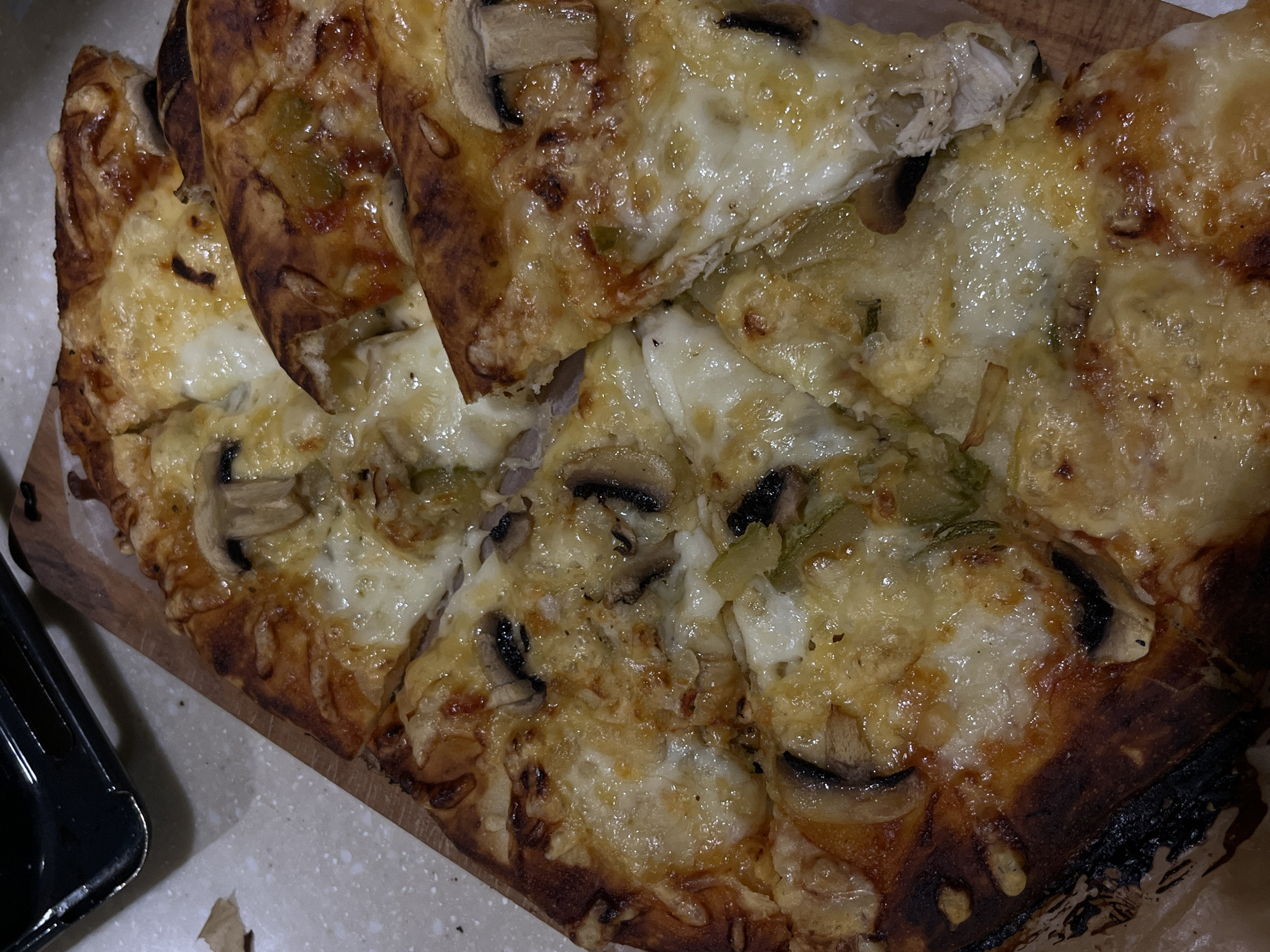 A close-up, overhead shot shows a whole pizza cut into slices on a wooden board. The pizza has a golden-brown crust and is topped with melted cheese, mushrooms, and green peppers. The pizza is slightly charred in places. A black container is visible in the upper left corner of the image, and a piece of brown paper is in the lower left. The background is a speckled, light-colored surface. The image is taken in Koroviia, Ukraine.
