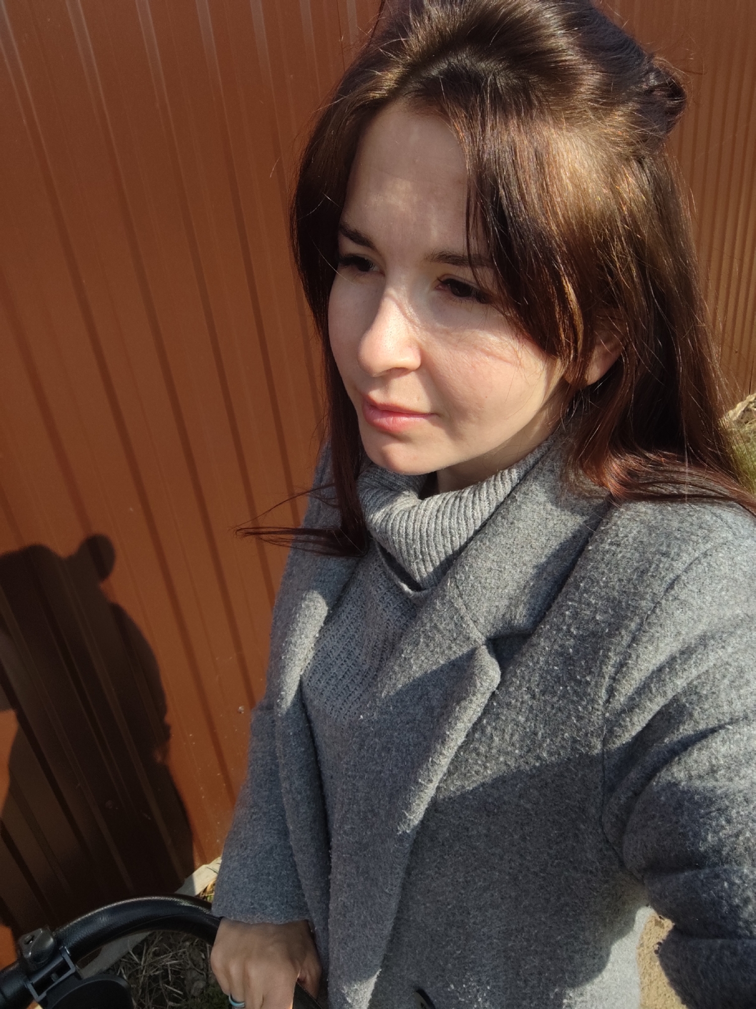 A young woman with dark brown hair, styled with some of it pulled back, stands outdoors wearing a grey turtleneck sweater and a grey coat. She's holding a black handlebar, possibly of a bicycle or stroller, in her hand. The backdrop is a brown corrugated metal fence. A shadow, likely cast by the person taking the photo, is visible on the fence. The location is likely the city of Фоки, Russia, due to the image's origin.