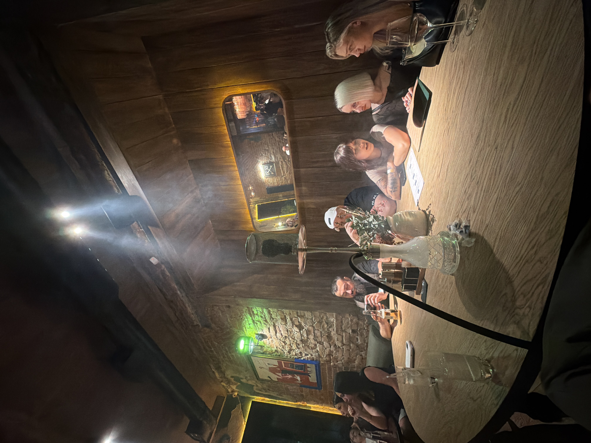 A group of six people are seated around a round wooden table in what appears to be a dimly lit hookah lounge. The individuals, appearing to be in their late 20s to early 40s, are engaged in conversation and drinking. The environment is characterized by exposed brick walls, wood paneling, and atmospheric lighting, with a prominent hookah pipe on the table. A painting hangs on the brick wall, and a mirror reflects parts of the room. The image captures a moment of social interaction in a relaxed, intimate setting. Based on the visual cues of the interior design and the atmosphere, this establishment is likely a modern lounge or bar in Krasnoyarsk, Russia, offering a unique ambiance for patrons.