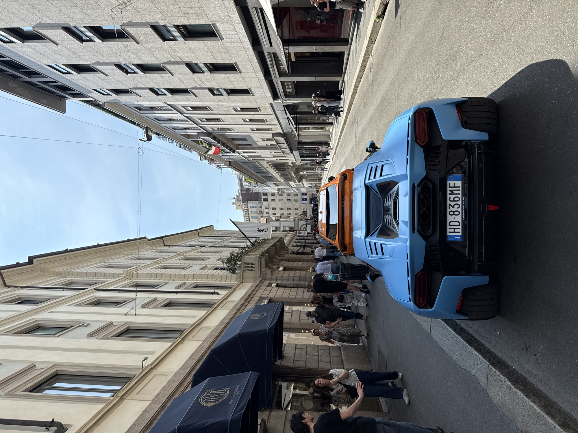 This image shows a street in Milan, Italy, lined with luxury boutiques, many with awnings and displays. People are walking on the sidewalks along the street. Two Lamborghinis, one light blue and the other orange, are parked along the side of the street. The light blue vehicle is facing away from the camera, showing its Italian license plate. Italian flags are visible further down the street. The buildings on either side of the street are tall, with many windows, and appear to be high-end retail spaces.