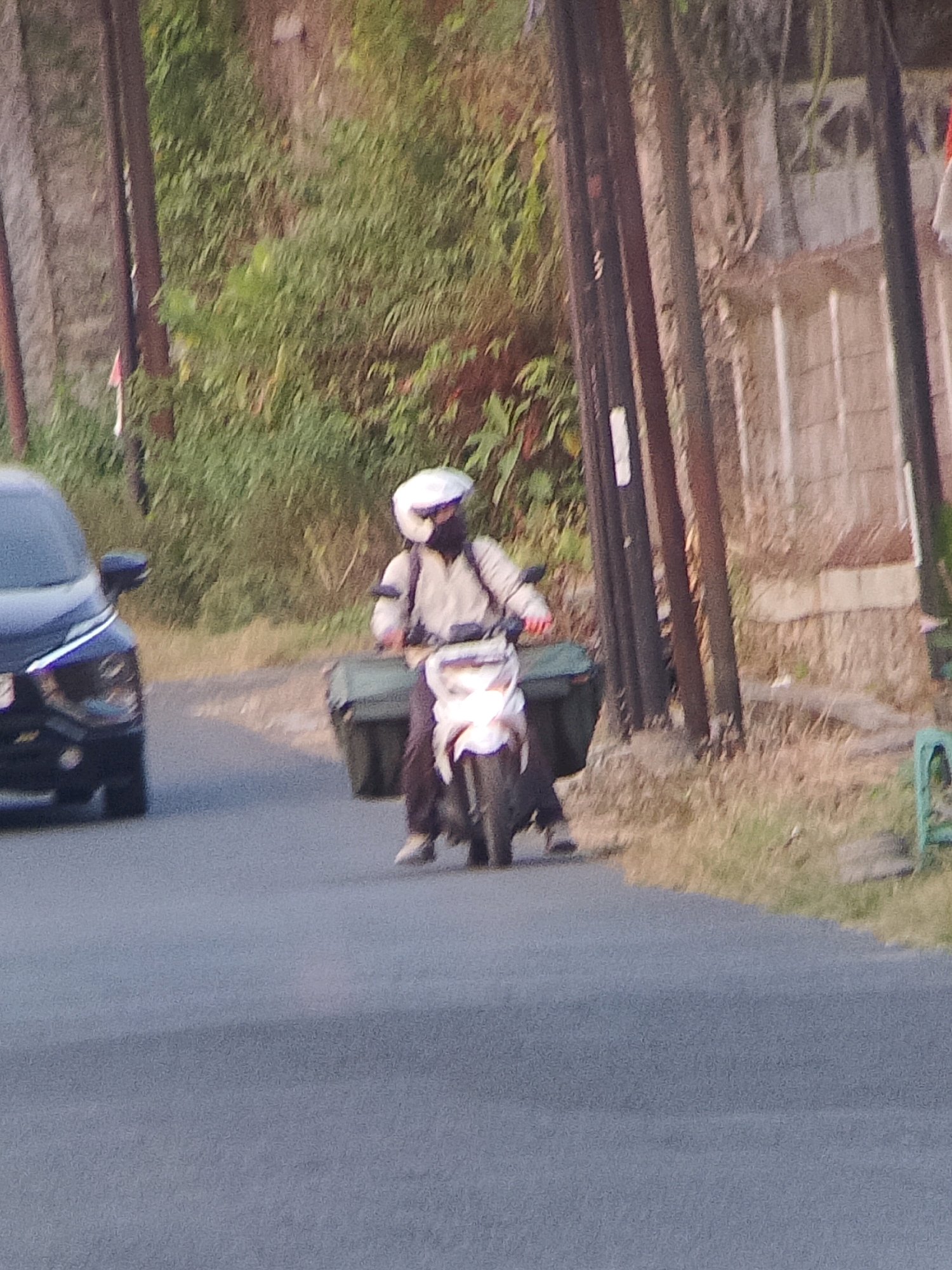 An individual wearing a white helmet and a dark mask is riding a white scooter along a paved road. The rider is dressed in a light-colored jacket and dark pants, and is carrying two large green bags attached to the rear of the scooter. To the left of the scooter, a dark-colored car is visible, partially obscuring the view of the rider and scooter. The background consists of a dense green area of trees and foliage, with some tall, dark poles interspersed throughout. A stone wall is visible on the far right, with a section of what appears to be a building behind it. The scene suggests a typical street in Jember, Indonesia, likely in a residential or semi-rural area, given the presence of both vehicles and abundant vegetation.