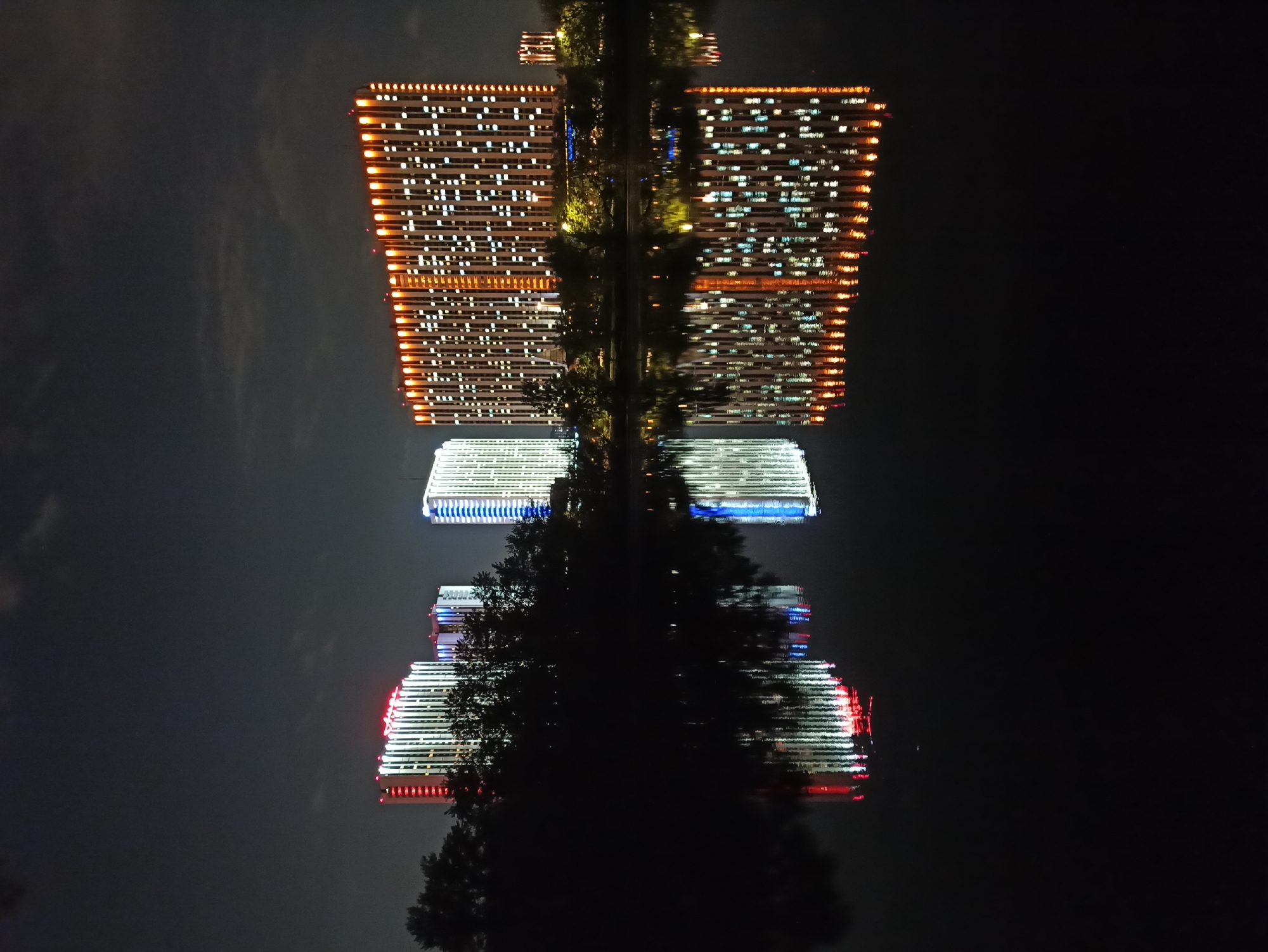 The image shows a nighttime reflection of buildings in water. The buildings appear to be modern office structures with many illuminated windows. There's a distinct pattern of orange, white, blue, and red lights on the buildings. Trees are interspersed between the buildings and their reflection. The location may be in Moscow, Russia.