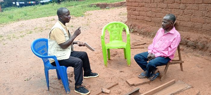 This image depicts an outdoor scene in Ngalula, Democratic Republic of the Congo, featuring two adult males engaged in an interaction. The ground is composed of reddish-brown earth. In the foreground, scattered pieces of unfinished wooden planks are present.
On the left, a male, appearing middle-aged, is seated on a blue plastic chair. He wears a light-colored short-sleeved shirt, a multi-pocketed light-colored vest, dark trousers, and dark sneakers. He holds a white rectangular object, possibly a tablet or clipboard, in his left hand and gestures with his raised right index finger, directing his gaze towards the other individual.
Between the two individuals, an unoccupied green plastic chair is visible.
On the right, an older male is seated on a small, three-legged wooden stool. He wears a pink long-sleeved button-up shirt, blue jeans, and blue sandals. His hands are clasped in his lap, and his gaze is directed to his left, slightly away from the first individual. Behind him, a wall constructed from reddish-brown mud bricks forms the immediate background.
Further in the background, to the left, green vegetation and a dirt path are visible, with what appears to be laundry hanging on a line in the distance.