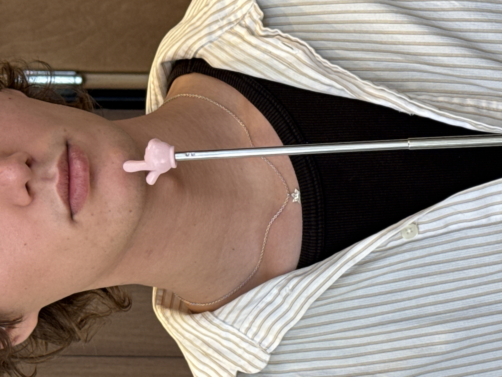 A close-up shot shows a person lying down, likely indoors given the flooring visible in the background. The person is wearing a horizontally striped collared shirt over a black top, and a delicate chain necklace. A silver stick is held, with a pink hand-shaped object on the end, pointing towards the person's face near their mouth. The context of the setting in Tel-Aviv, Israel is not evident from the image.