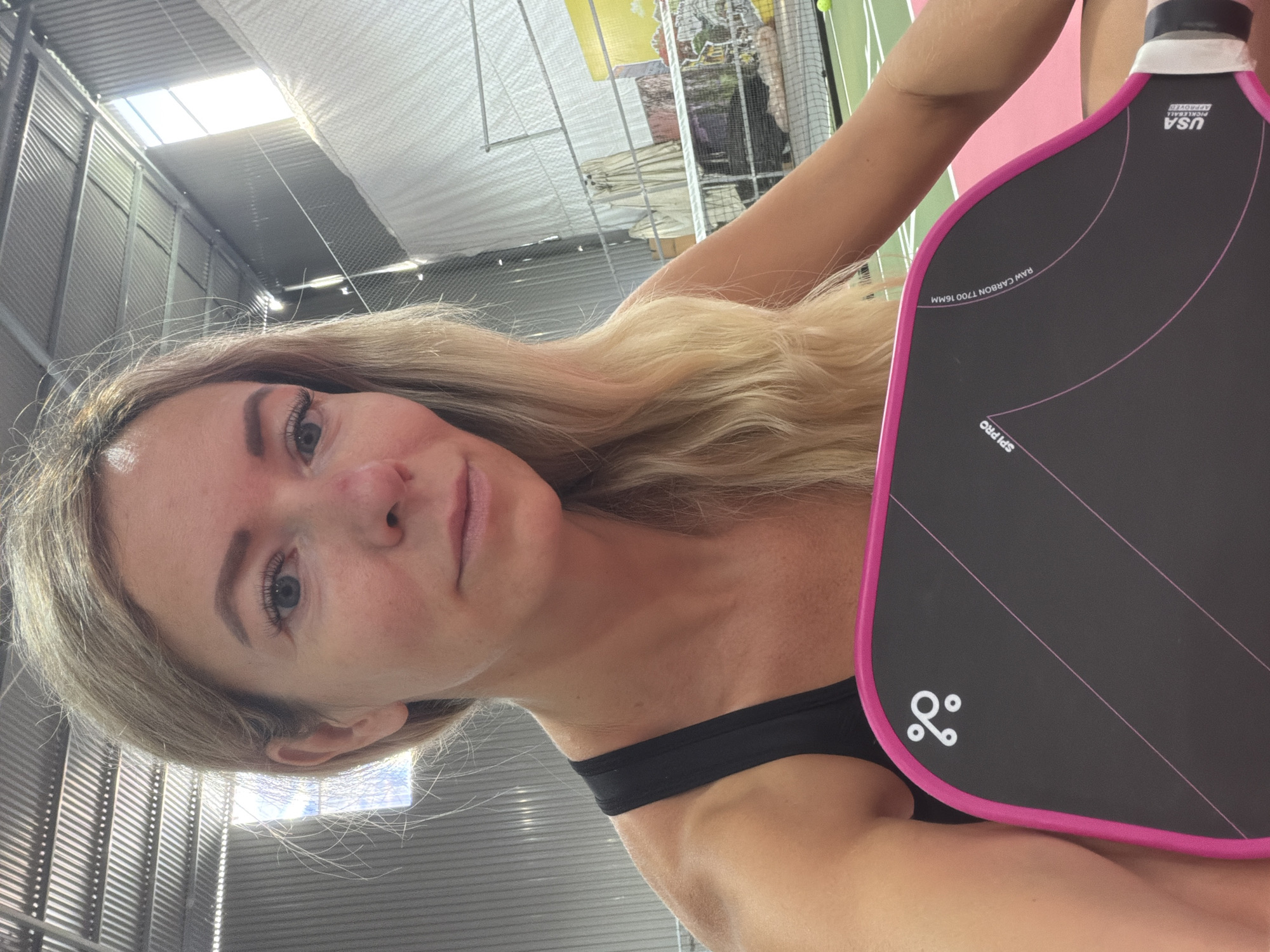 A woman with blonde hair is holding a pickleball paddle in an indoor pickleball court. She is wearing a black tank top and has a pink border on the paddle. The court appears to be empty and there are tennis nets and a court in the background. The location is identified as a pickleball court in Da Lat, Vietnam.
