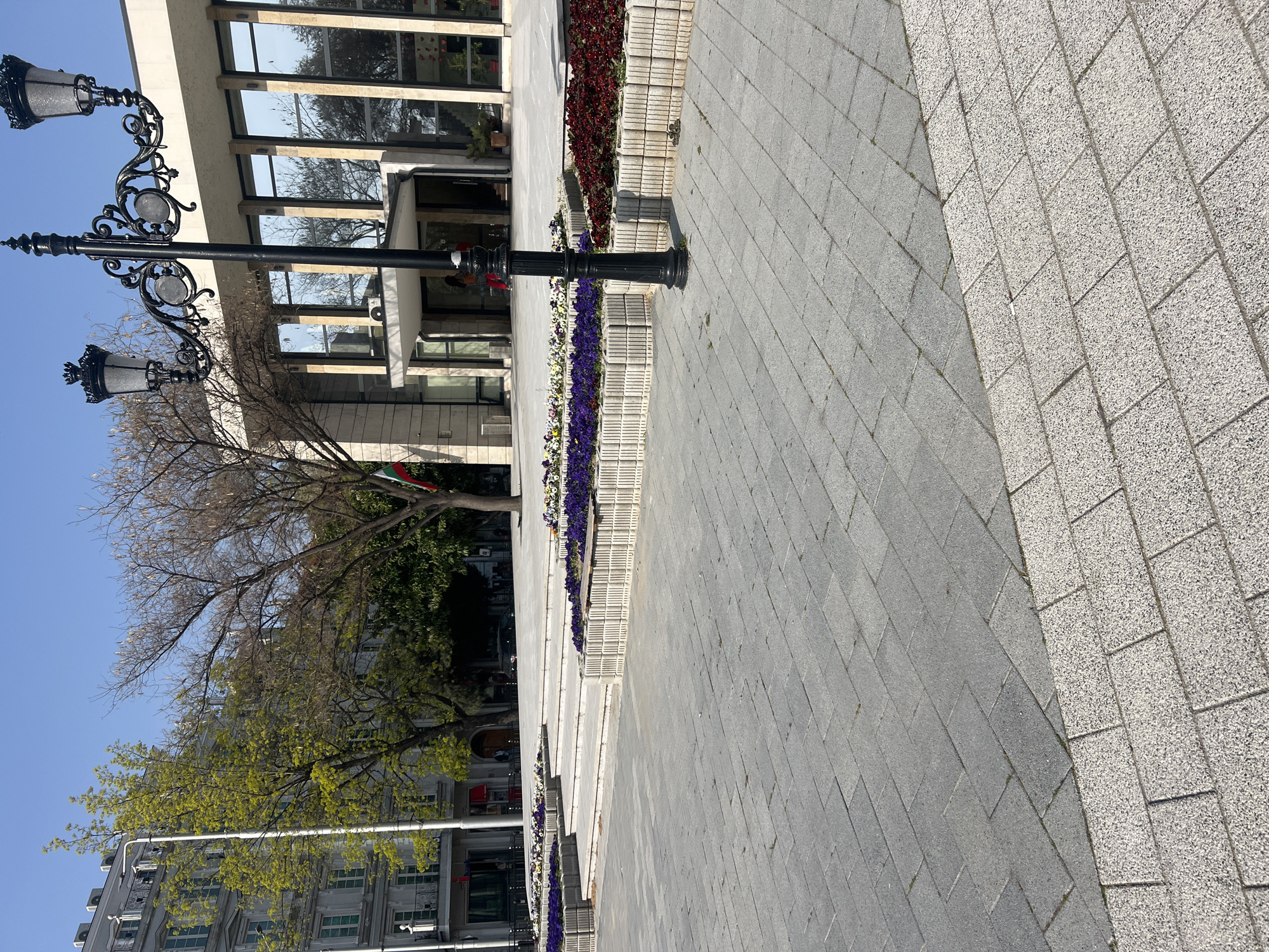The image captures a street scene in Plovdiv, Bulgaria. In the foreground, there is a paved pedestrian walkway, with a distinct change in paving texture. Adjacent to the walkway is a tiered planter with flowers. A black, ornate street lamp stands on the left side of the frame. Behind the lamp and planters is a modern building with glass windows reflecting trees and sky. Trees with varying degrees of foliage are visible, suggesting different stages of the spring season. In the background, parts of another building can be seen, suggesting an urban setting. The sky is clear and blue. No people are clearly visible in the image.